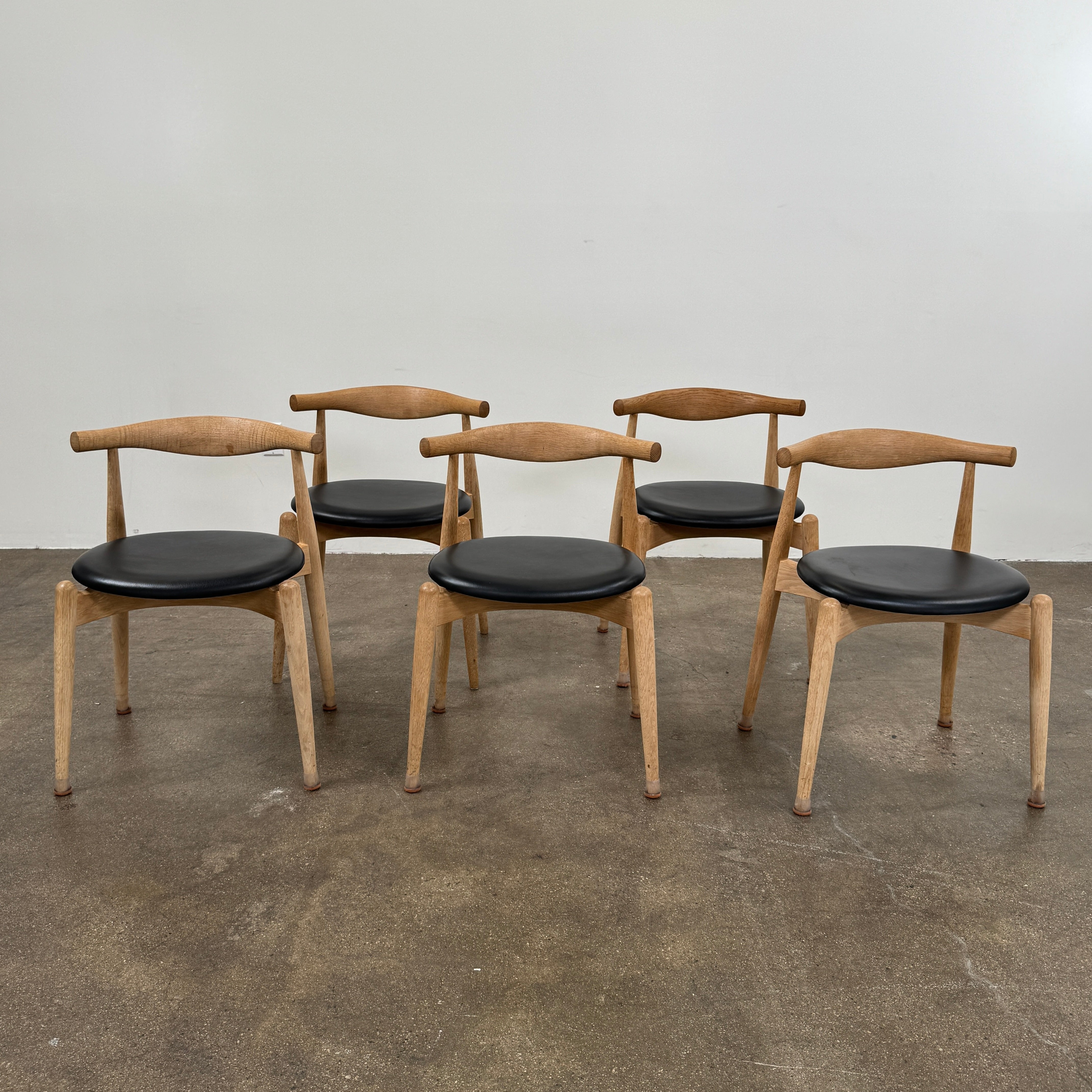 A set of five Carl Hansen & Son CH20 Elbow Chairs by Carl Hansen & Son, each with a curved wooden backrest and a round black seat cushion, are arranged in two rows against a plain white background.