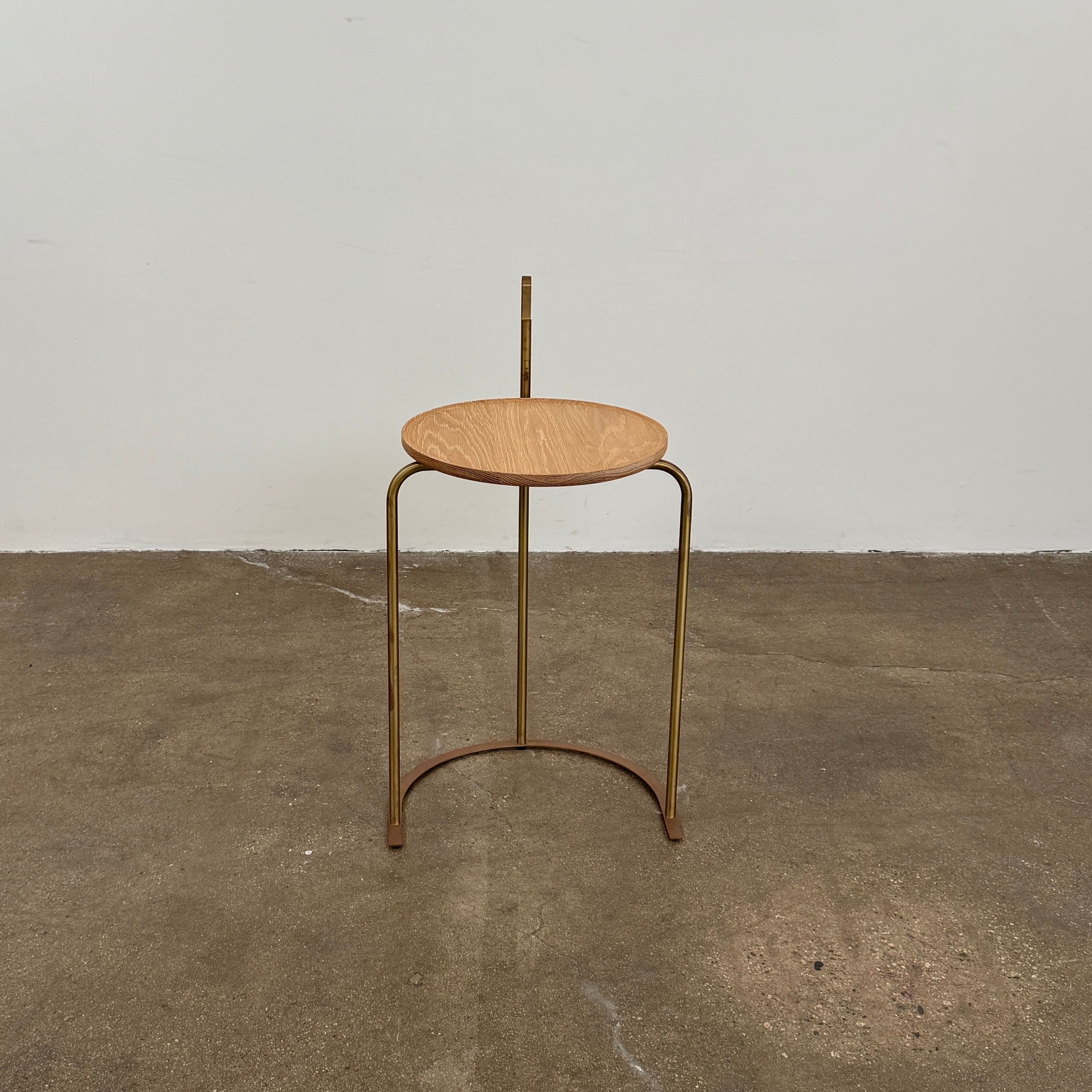 The De La Espada Handle Side Table by De La Espada features a small, round oiled oak top and a minimalist brass frame with a distinctive upright handle-like detail on one side.