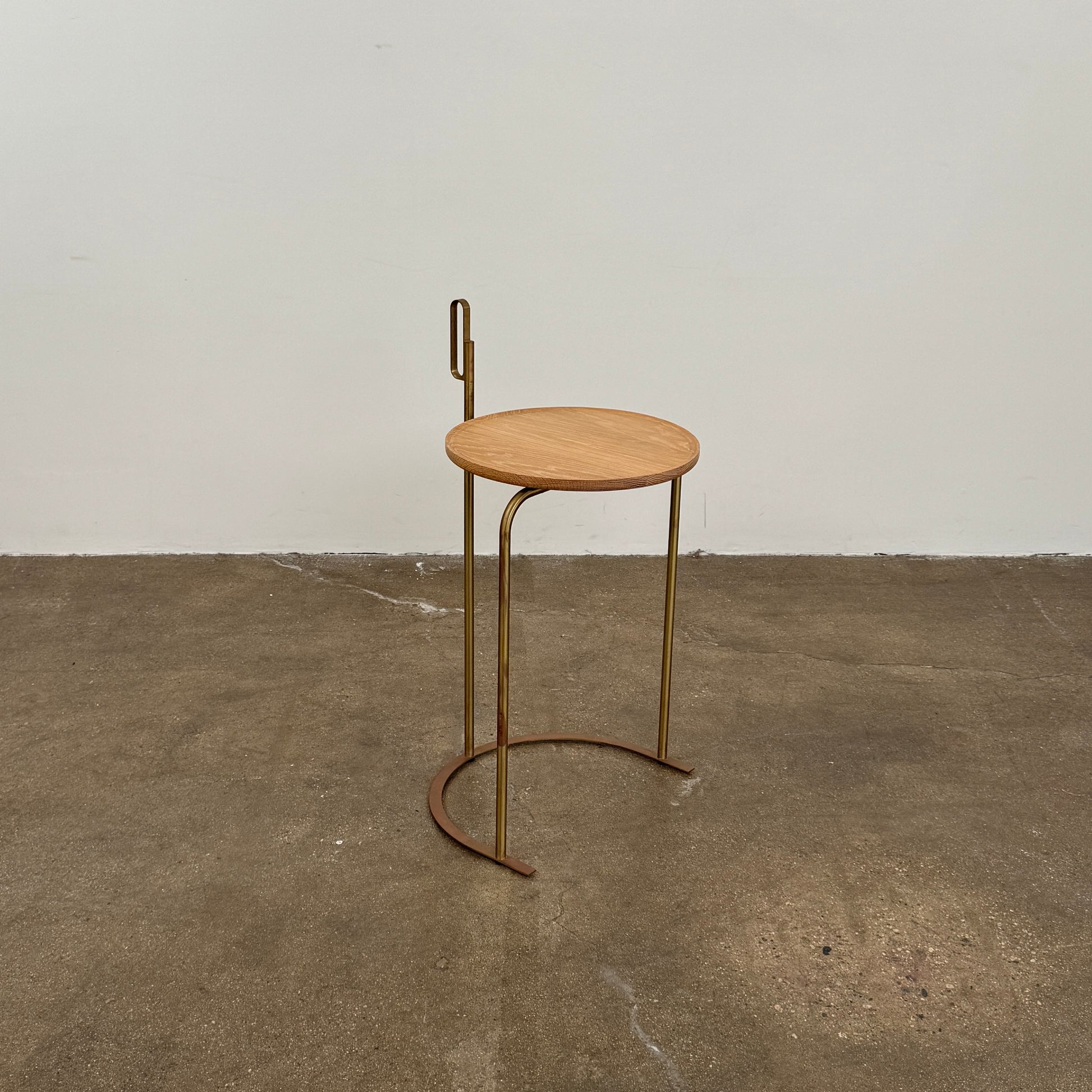 The De La Espada Handle Side Table by De La Espada features a small, round oiled oak top and a minimalist brass frame with a distinctive upright handle-like detail on one side.