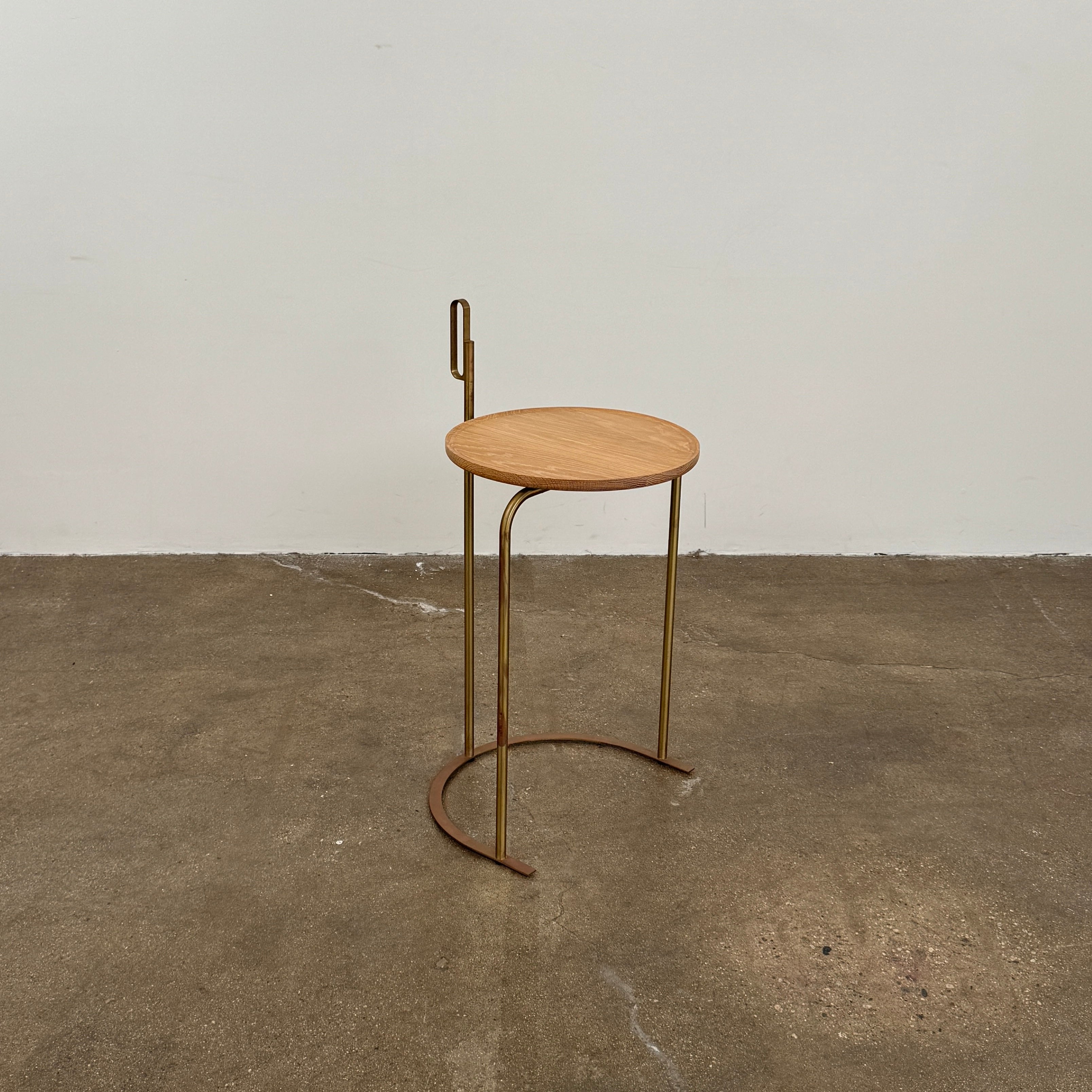 The De La Espada Handle Side Table by De La Espada features a small, round oiled oak top and a minimalist brass frame with a distinctive upright handle-like detail on one side.
