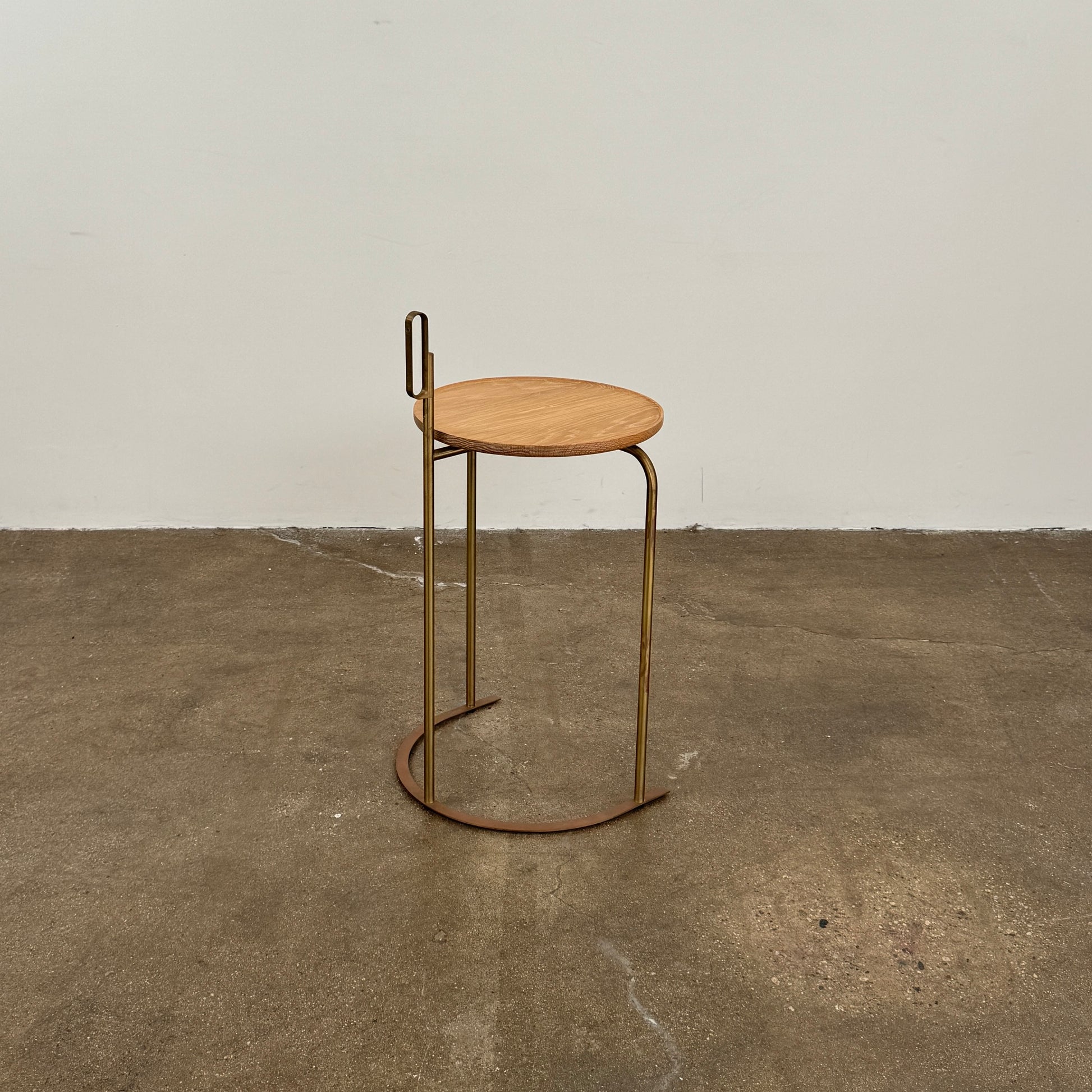 The De La Espada Handle Side Table by De La Espada features a small, round oiled oak top and a minimalist brass frame with a distinctive upright handle-like detail on one side.