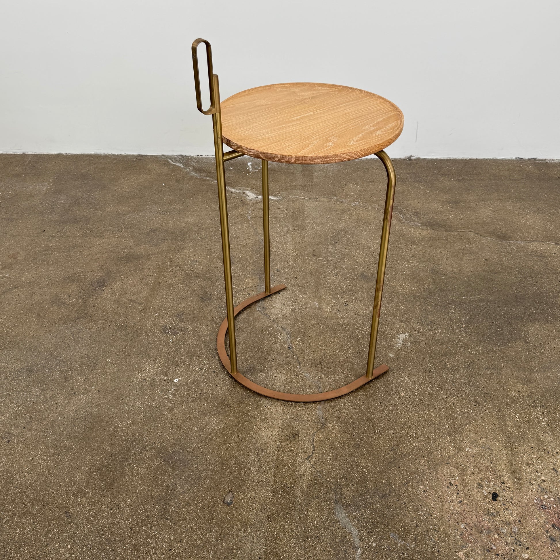 The De La Espada Handle Side Table by De La Espada features a small, round oiled oak top and a minimalist brass frame with a distinctive upright handle-like detail on one side.