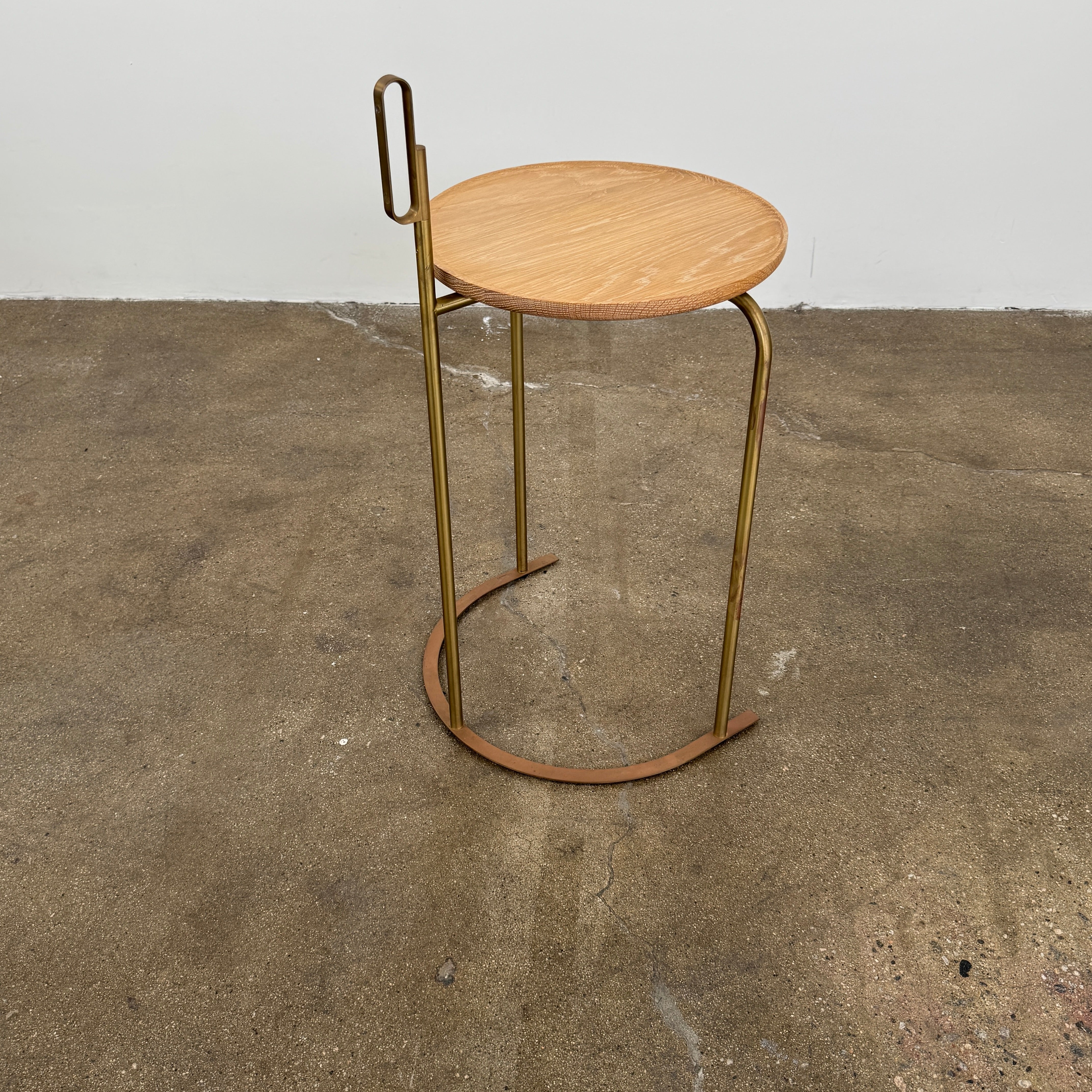 The De La Espada Handle Side Table by De La Espada features a small, round oiled oak top and a minimalist brass frame with a distinctive upright handle-like detail on one side.