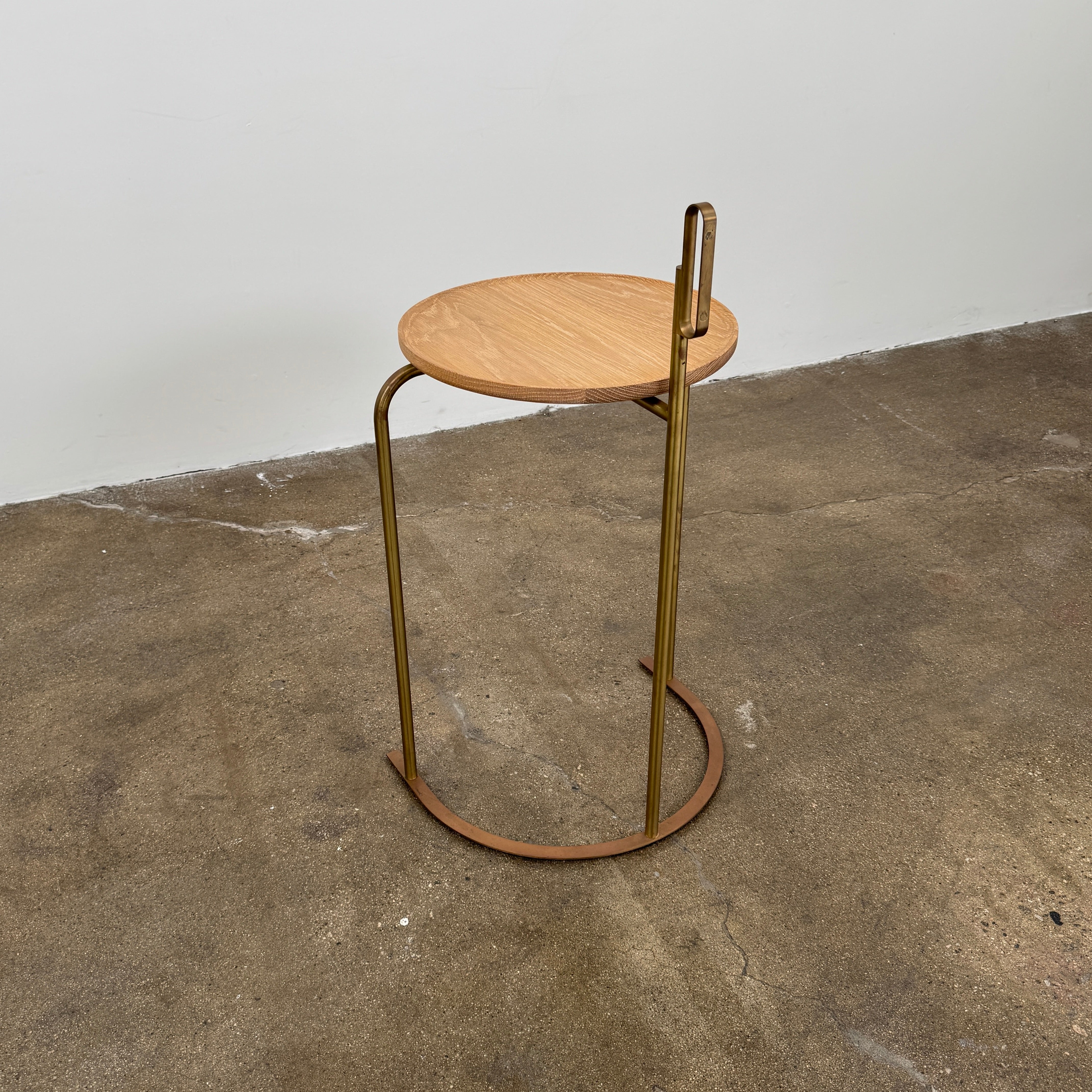 The De La Espada Handle Side Table by De La Espada features a small, round oiled oak top and a minimalist brass frame with a distinctive upright handle-like detail on one side.