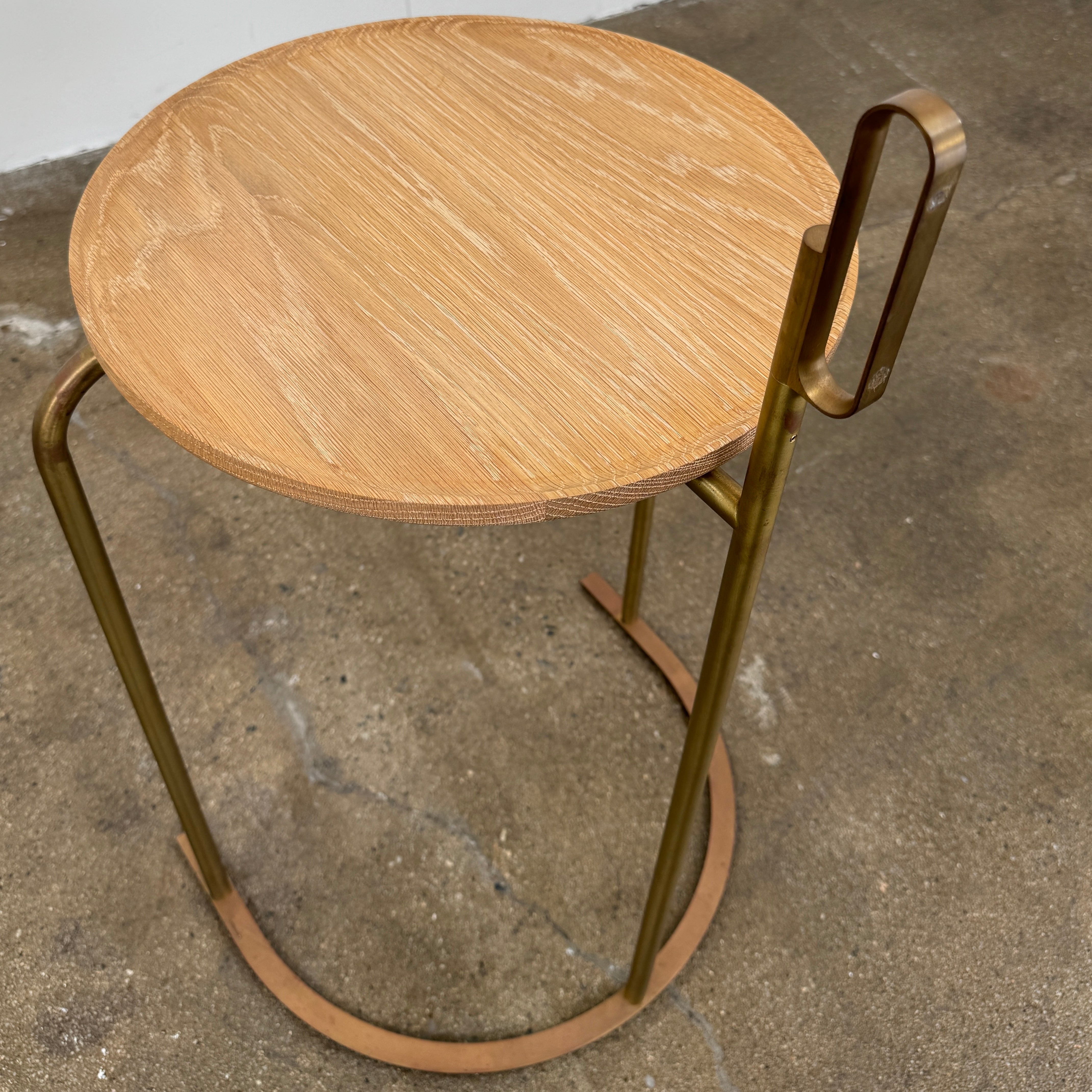 The De La Espada Handle Side Table by De La Espada features a small, round oiled oak top and a minimalist brass frame with a distinctive upright handle-like detail on one side.