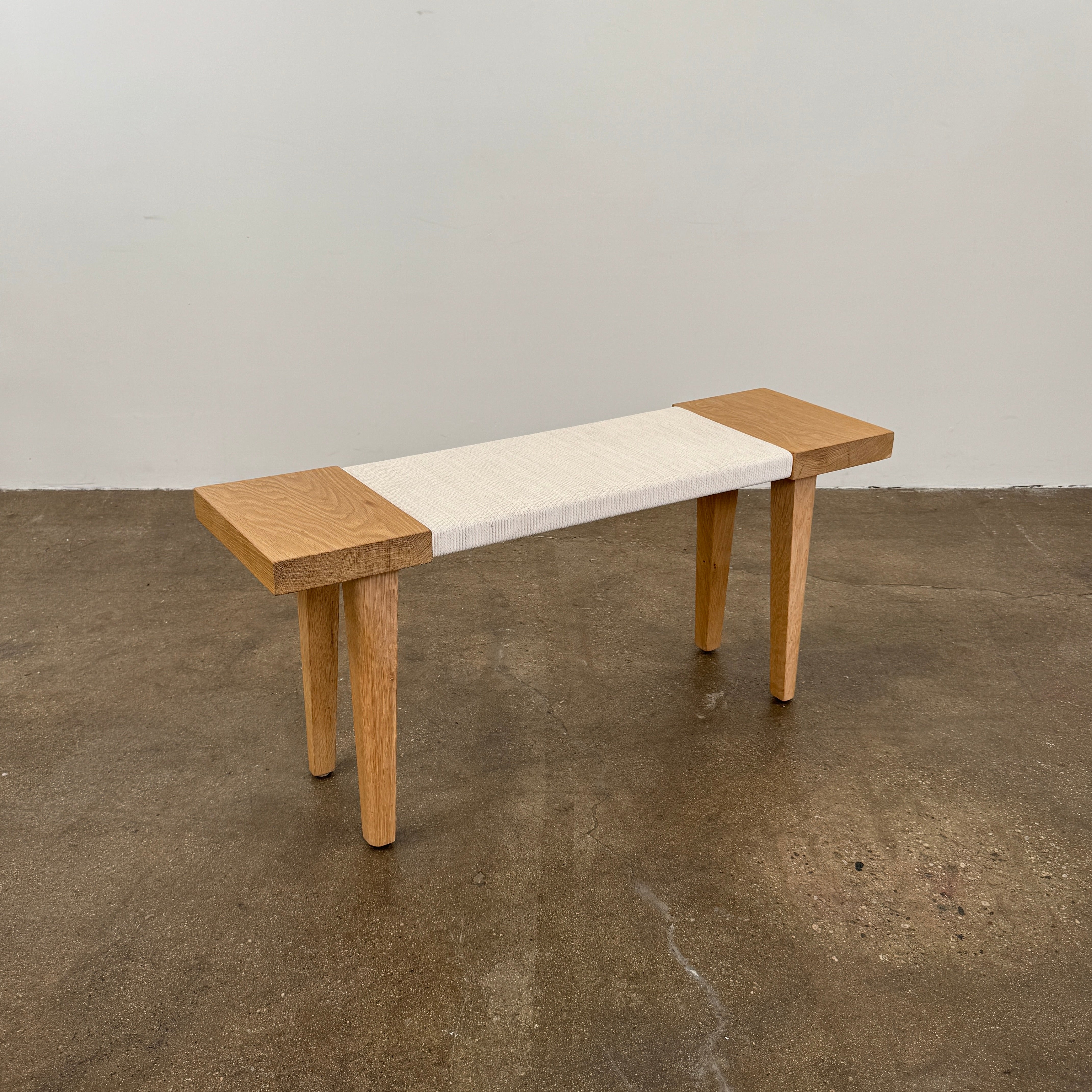 The Jenni Kayne Oak Day Bench by Jenni Kayne is a handcrafted White Oak piece with a minimalist design, featuring four legs and a light-colored woven seat, shown here on a polished concrete floor—an elegant addition to any Los Angeles furniture collection.