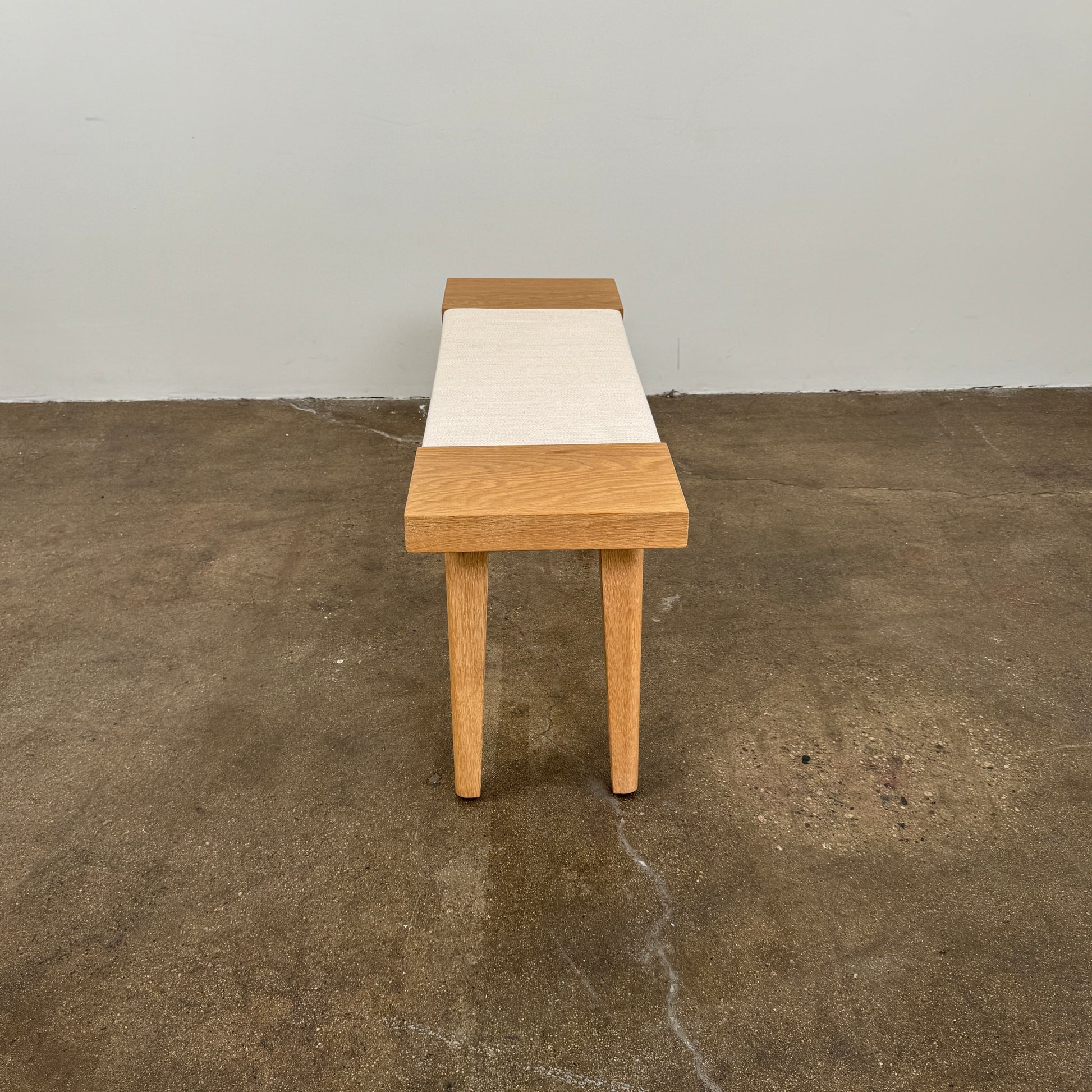 The Jenni Kayne Oak Day Bench by Jenni Kayne is a handcrafted White Oak piece with a minimalist design, featuring four legs and a light-colored woven seat, shown here on a polished concrete floor—an elegant addition to any Los Angeles furniture collection.