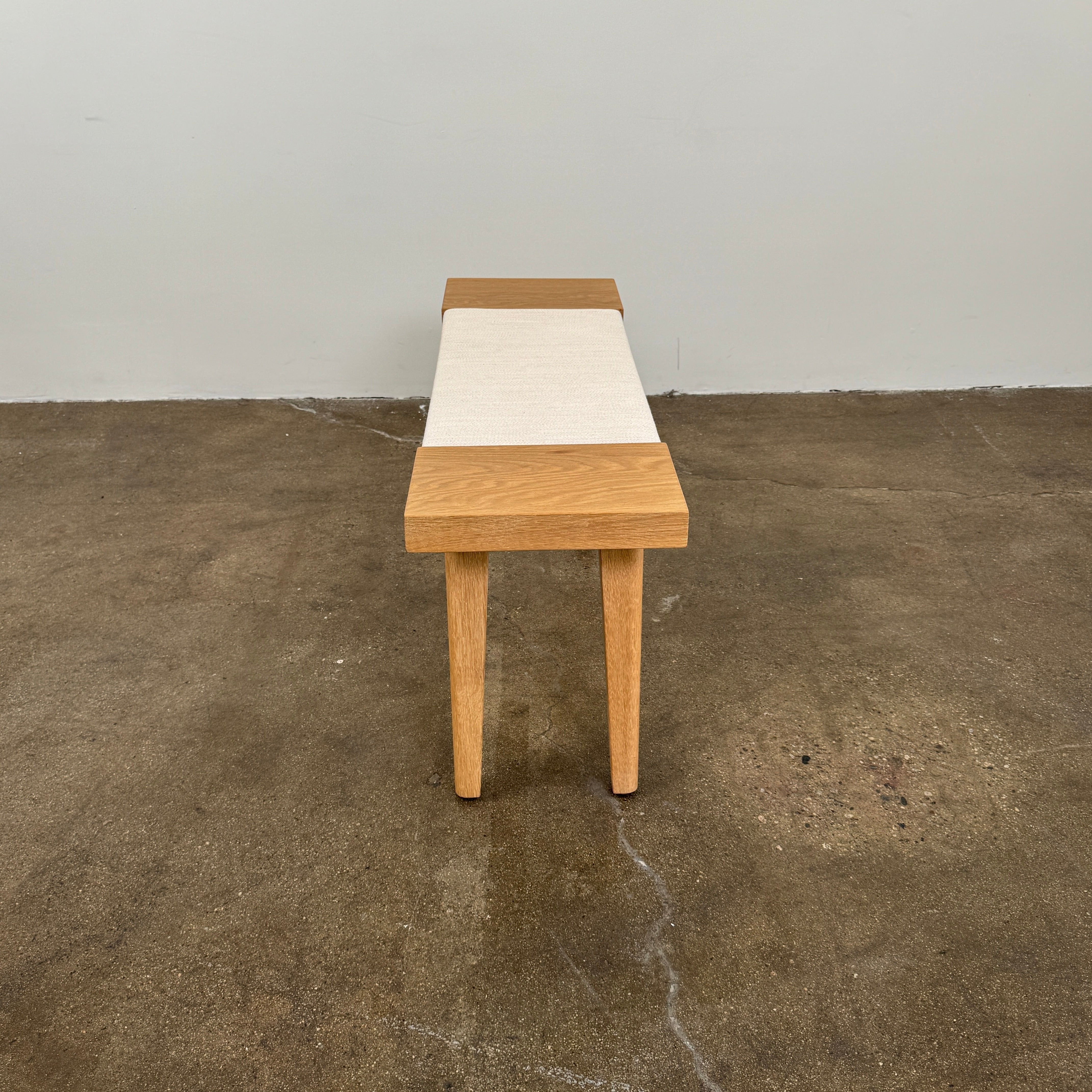 The Jenni Kayne Oak Day Bench by Jenni Kayne is a handcrafted White Oak piece with a minimalist design, featuring four legs and a light-colored woven seat, shown here on a polished concrete floor—an elegant addition to any Los Angeles furniture collection.