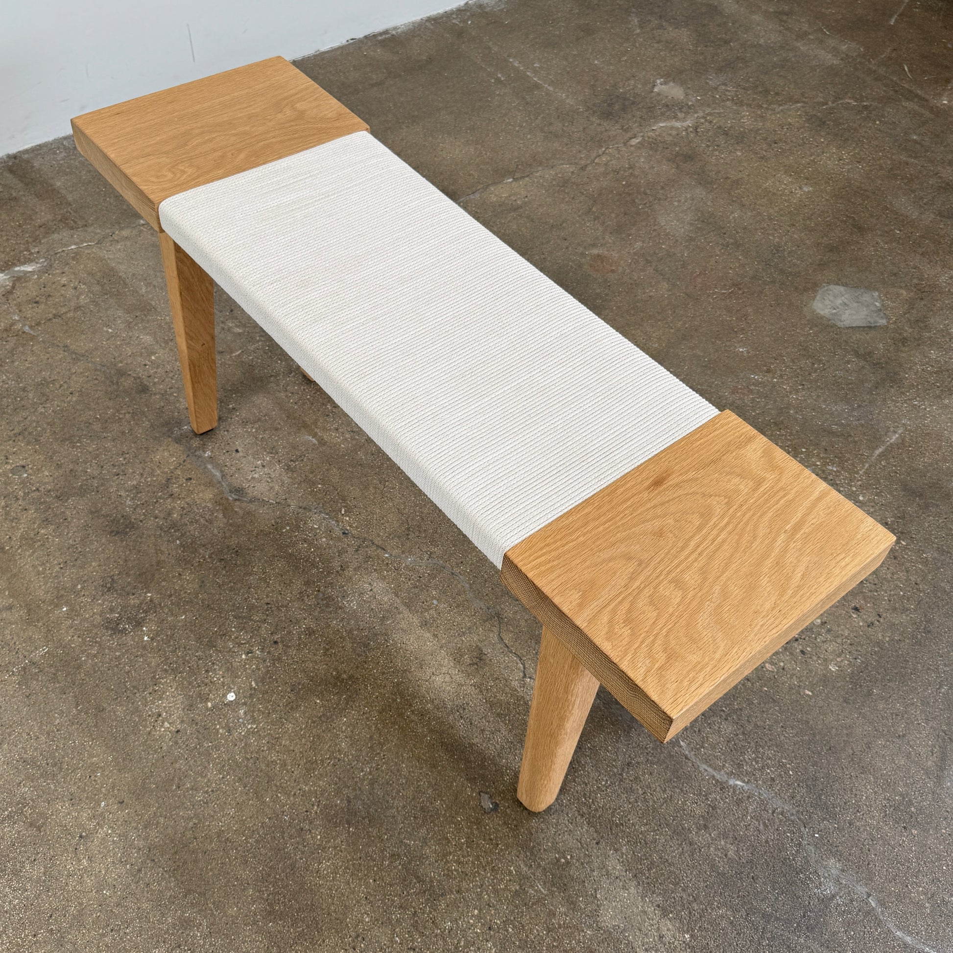 The Jenni Kayne Oak Day Bench by Jenni Kayne is a handcrafted White Oak piece with a minimalist design, featuring four legs and a light-colored woven seat, shown here on a polished concrete floor—an elegant addition to any Los Angeles furniture collection.