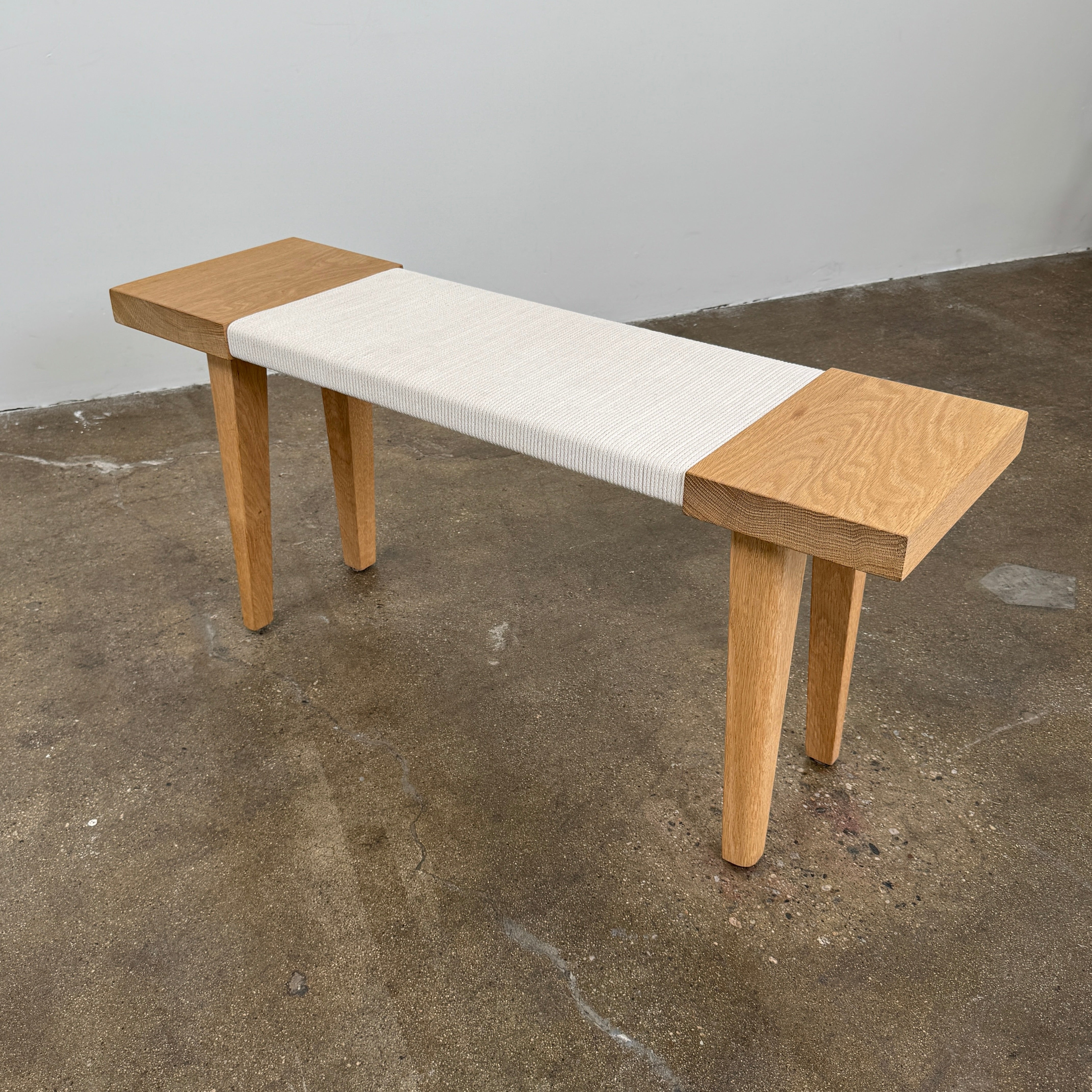 The Jenni Kayne Oak Day Bench by Jenni Kayne is a handcrafted White Oak piece with a minimalist design, featuring four legs and a light-colored woven seat, shown here on a polished concrete floor—an elegant addition to any Los Angeles furniture collection.