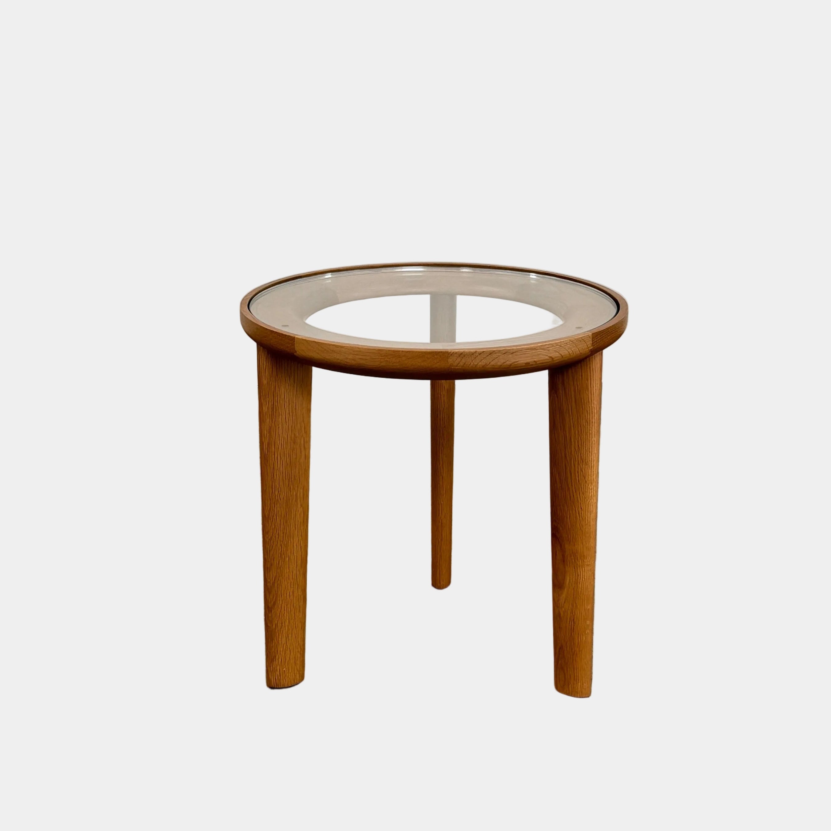 The DWR Port Side Table by DWR features a small, round design with three solid American oak legs and a clear glass top, shown against a plain white background.
