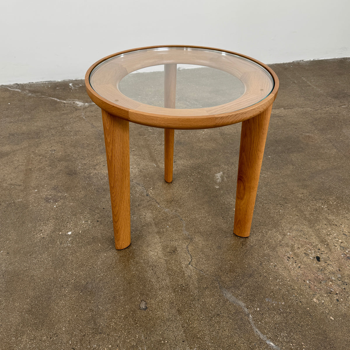 The DWR Port Side Table by DWR features a small, round design with three solid American oak legs and a clear glass top, shown against a plain white background.