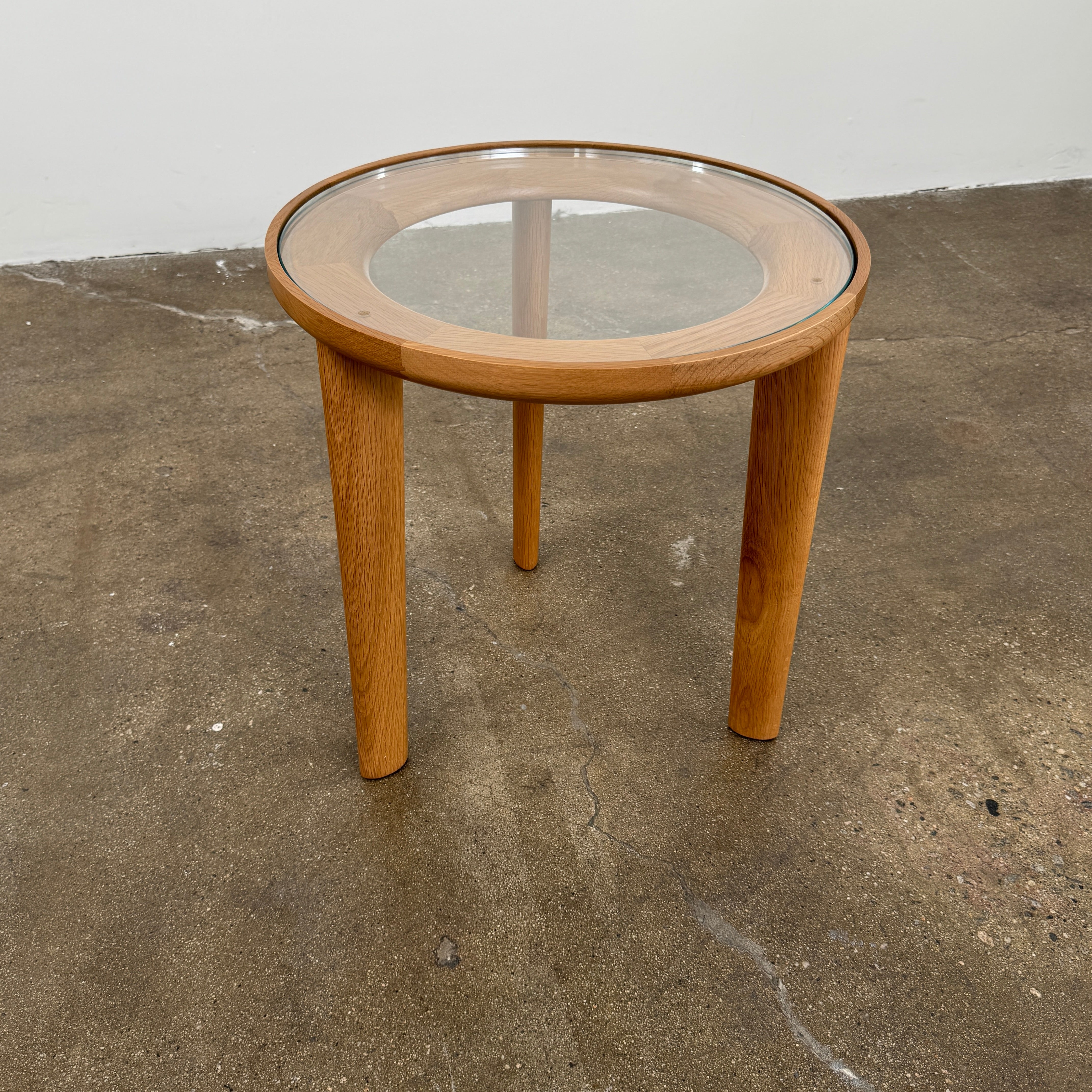 The DWR Port Side Table by DWR features a small, round design with three solid American oak legs and a clear glass top, shown against a plain white background.