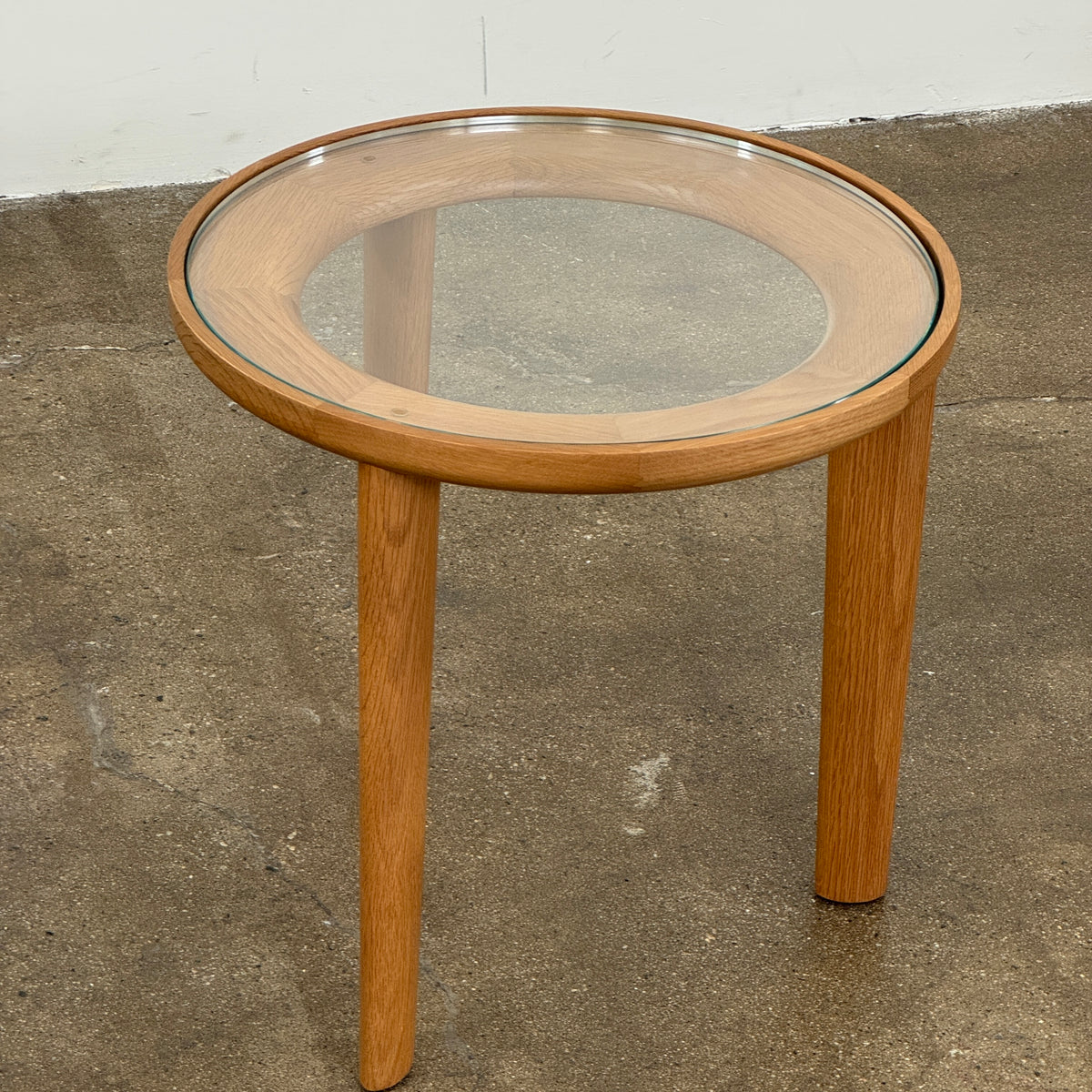 The DWR Port Side Table by DWR features a small, round design with three solid American oak legs and a clear glass top, shown against a plain white background.