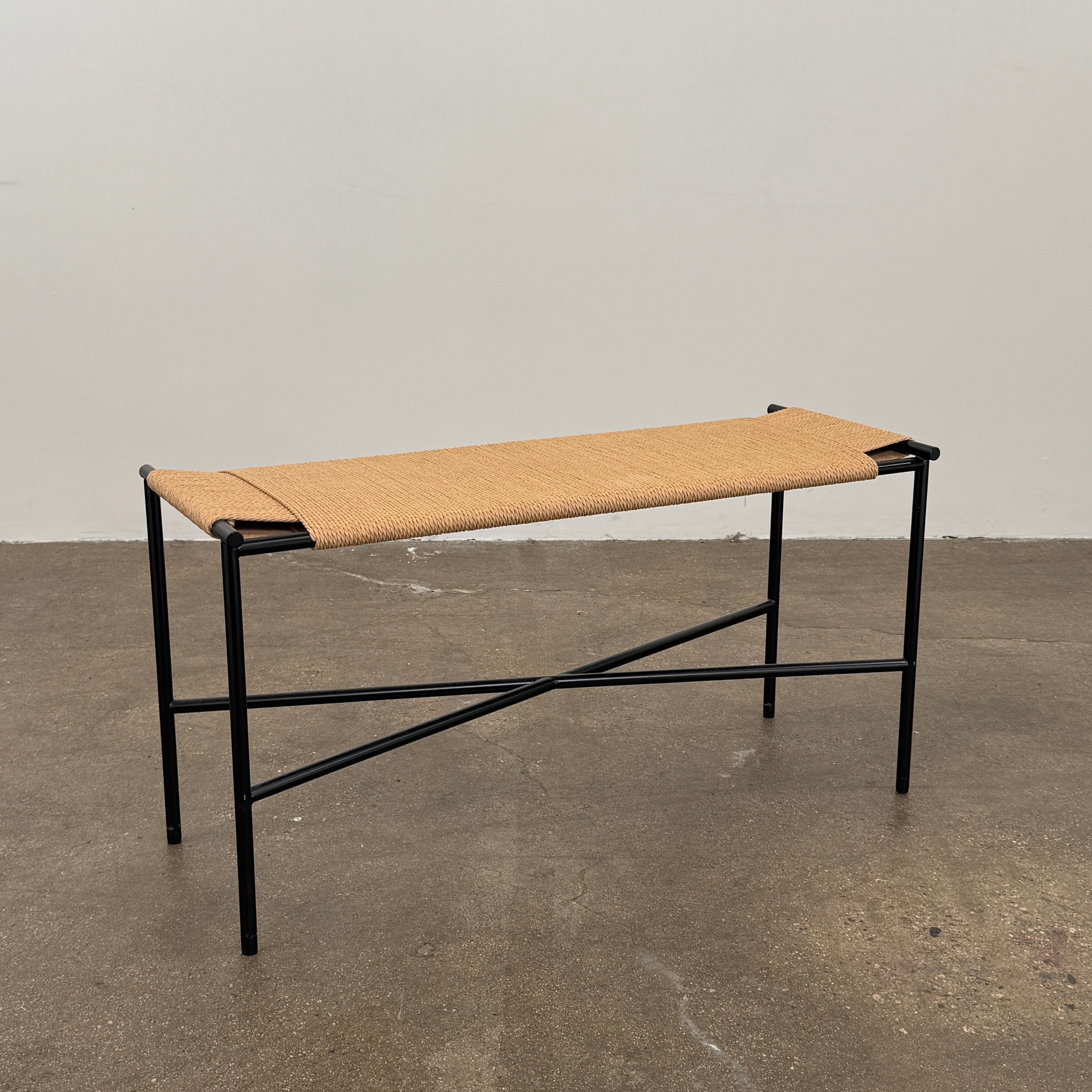 Fritz Hansen Skagerak VENT Bench by Fritz Hansen, featuring a rectangular black metal frame with minimalist crossed supports underneath and a woven tan paper cord seat, shown on a plain white background.