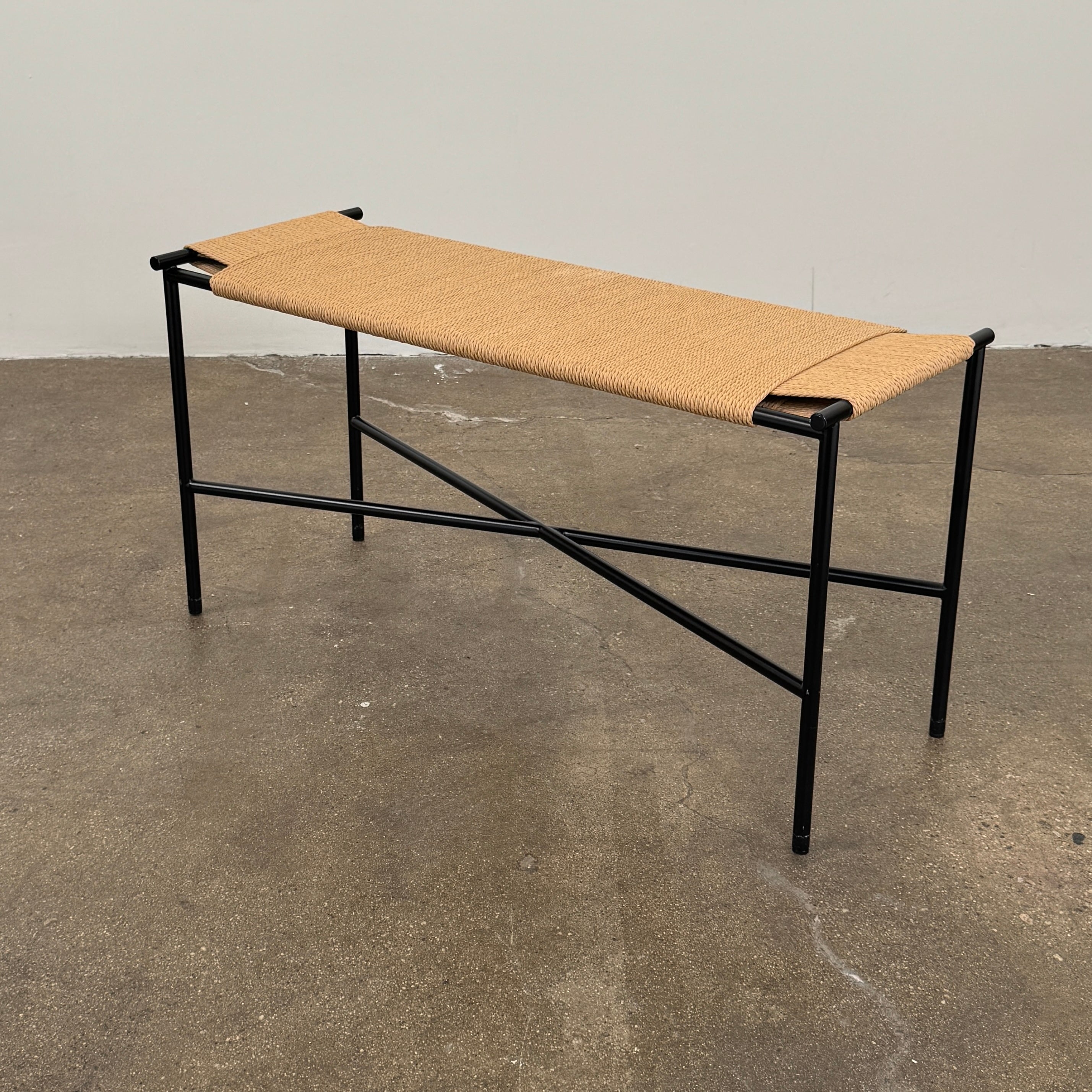 Fritz Hansen Skagerak VENT Bench by Fritz Hansen, featuring a rectangular black metal frame with minimalist crossed supports underneath and a woven tan paper cord seat, shown on a plain white background.