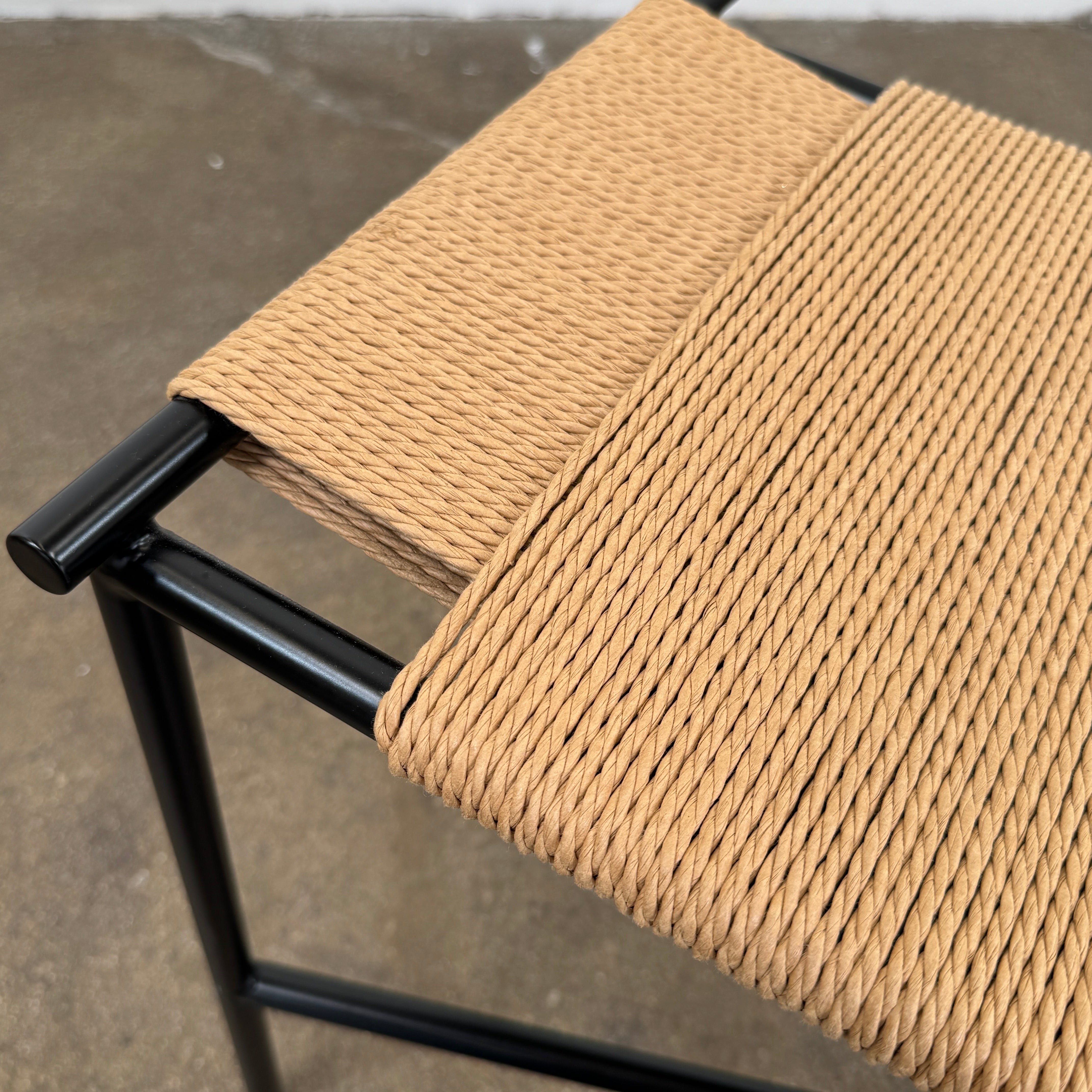Fritz Hansen Skagerak VENT Bench by Fritz Hansen, featuring a rectangular black metal frame with minimalist crossed supports underneath and a woven tan paper cord seat, shown on a plain white background.