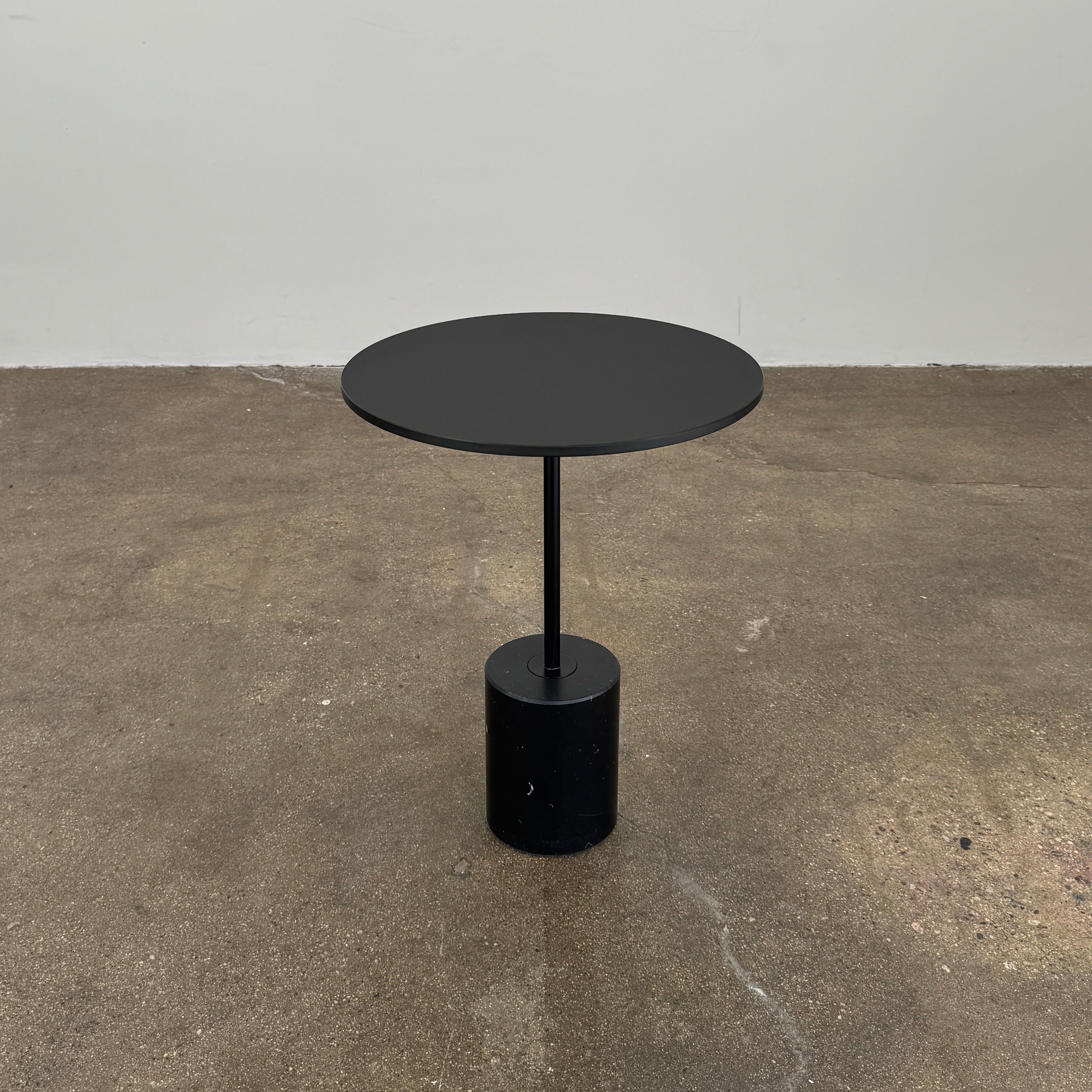La Palma’s Jey Side Table features a minimalist black round top supported by a slim central column and anchored with a solid marble cylinder base, all showcased against a plain light background.