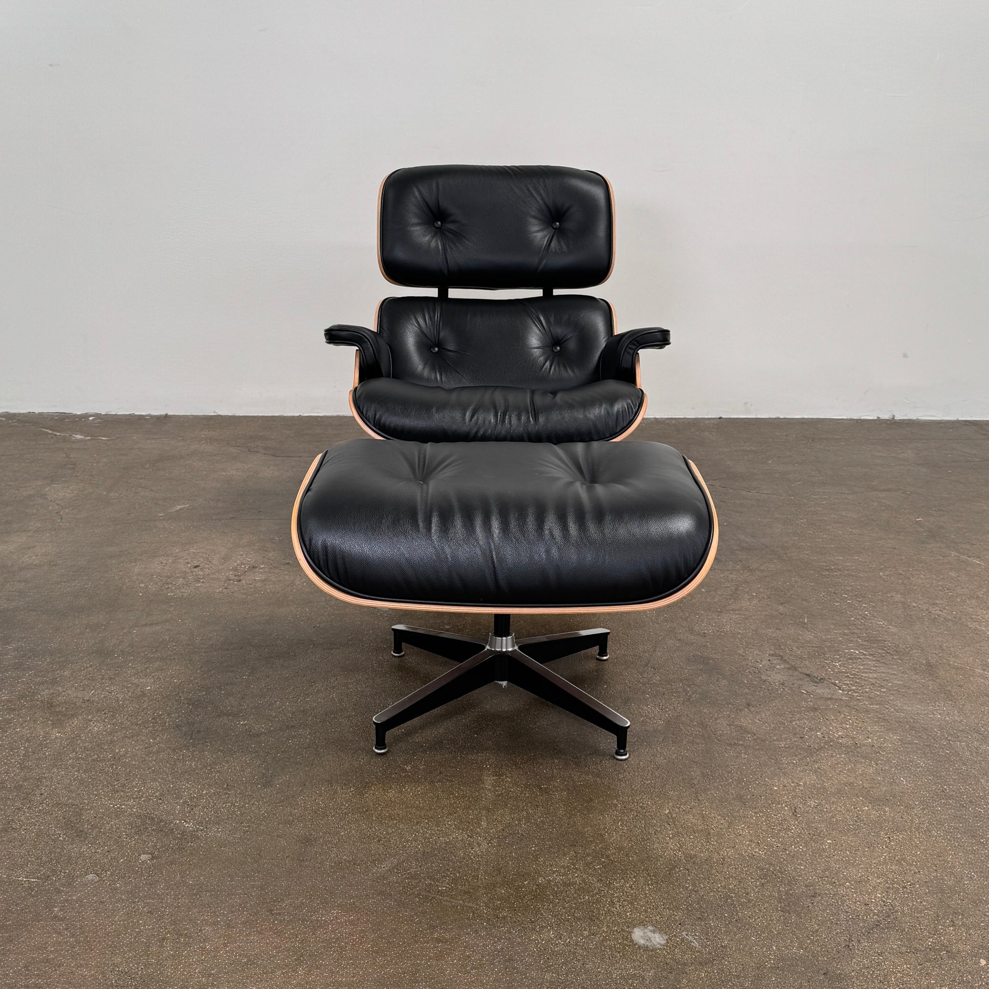 The Eames Lounge Chair and Ottoman by Modern Resale, upholstered in black leather with curved wooden accents, sits on a polished concrete floor against a plain light-colored wall.