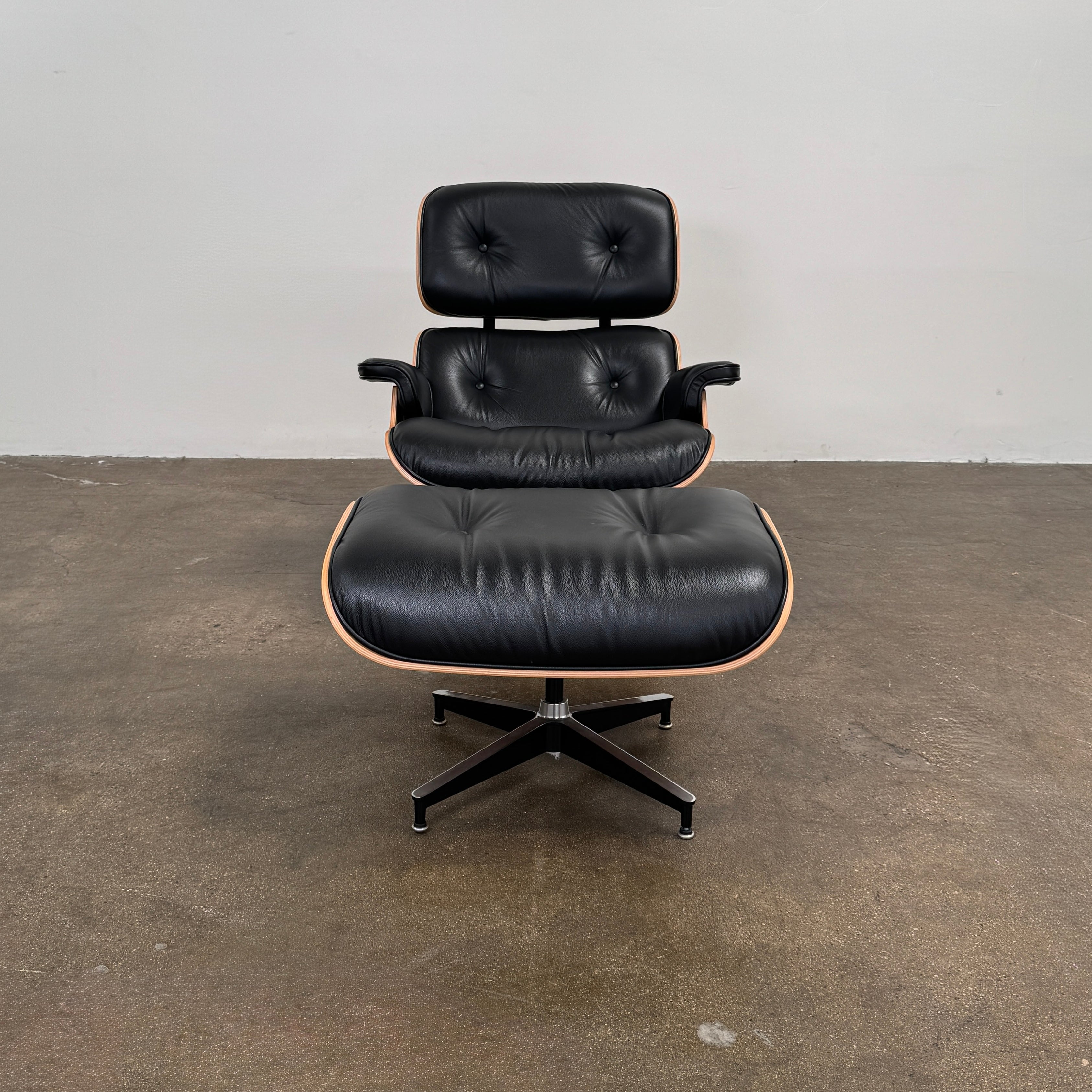 The Eames Lounge Chair and Ottoman by Modern Resale, upholstered in black leather with curved wooden accents, sits on a polished concrete floor against a plain light-colored wall.