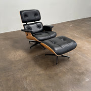The Eames Lounge Chair and Ottoman by Modern Resale, upholstered in black leather with curved wooden accents, sits on a polished concrete floor against a plain light-colored wall.