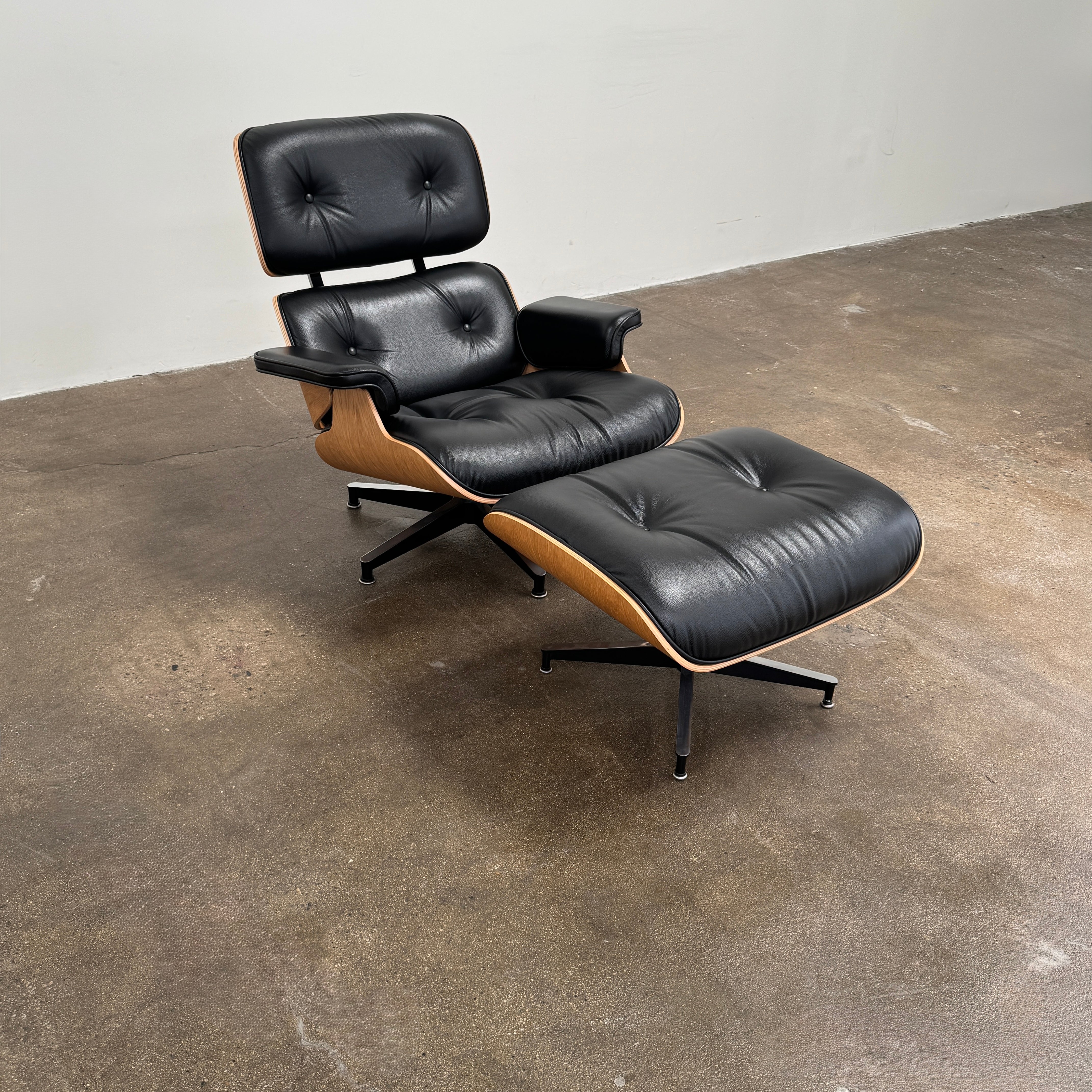 The Eames Lounge Chair and Ottoman by Modern Resale, upholstered in black leather with curved wooden accents, sits on a polished concrete floor against a plain light-colored wall.