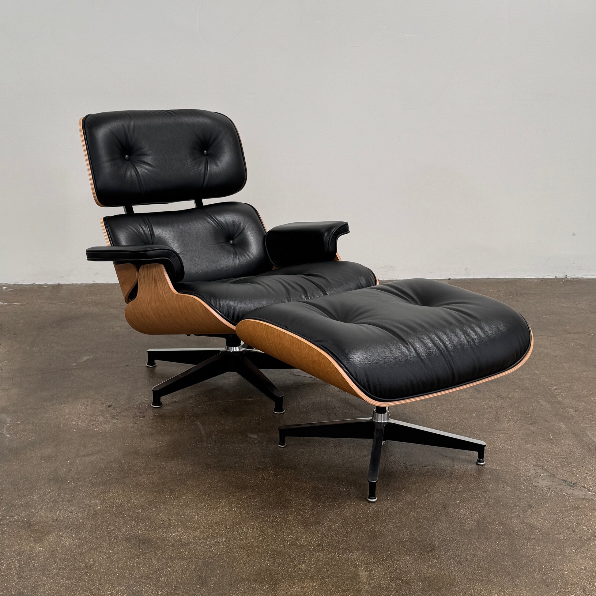 The Eames Lounge Chair and Ottoman by Modern Resale, upholstered in black leather with curved wooden accents, sits on a polished concrete floor against a plain light-colored wall.