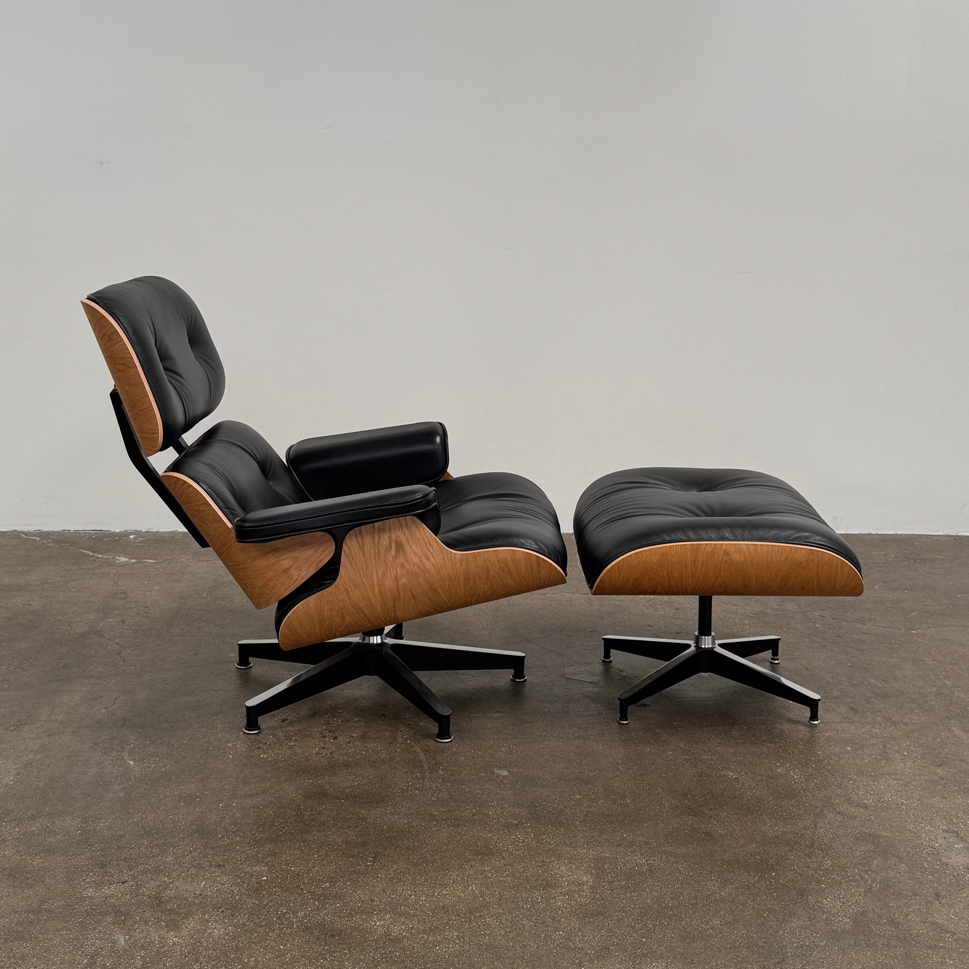 The Herman Miller Eames Lounge Chair & Ottoman (Copy) by Herman Miller, featuring black leather upholstery and modern design inspiration, is positioned on a brown concrete floor against a plain light-colored wall.