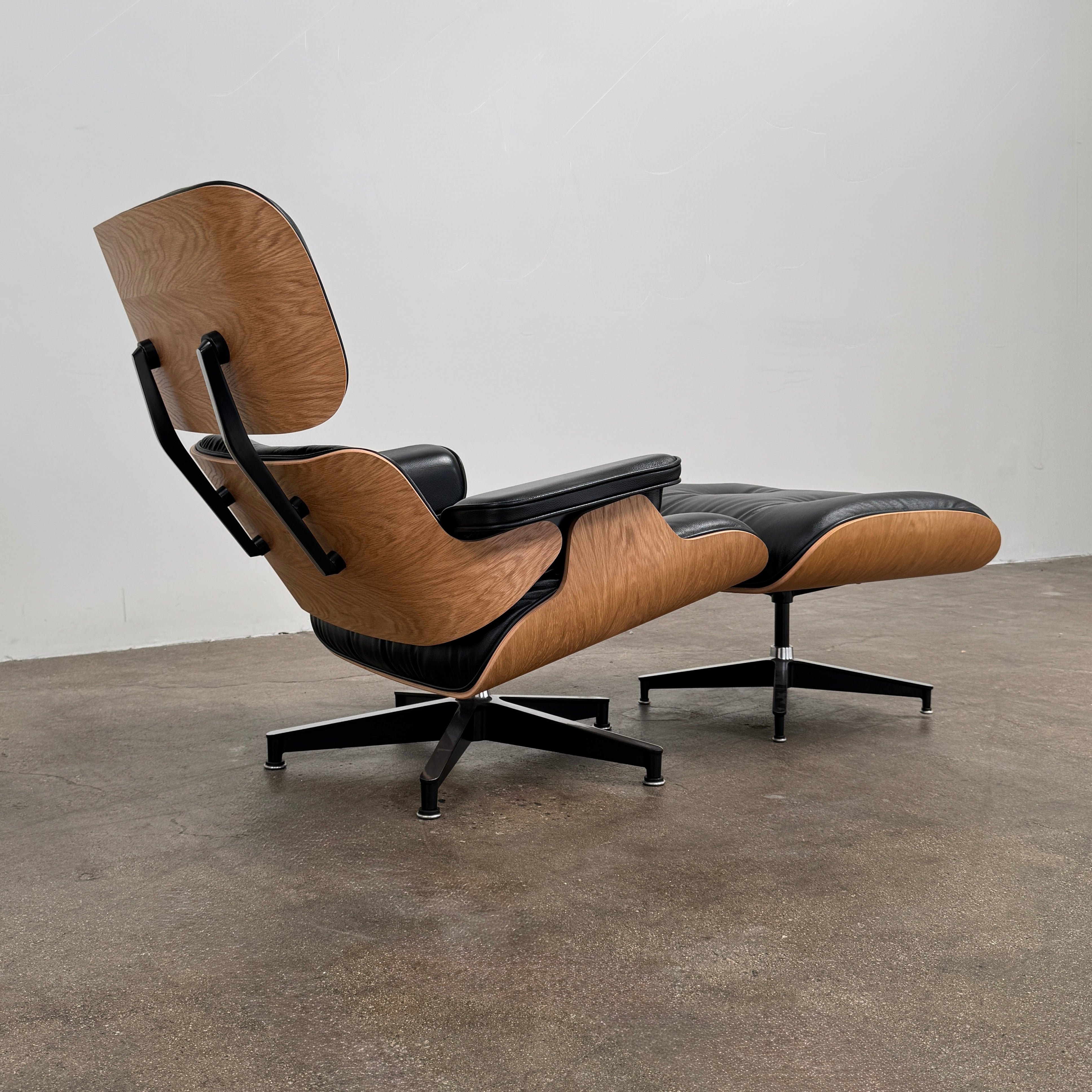 The Eames Lounge Chair and Ottoman by Modern Resale, upholstered in black leather with curved wooden accents, sits on a polished concrete floor against a plain light-colored wall.