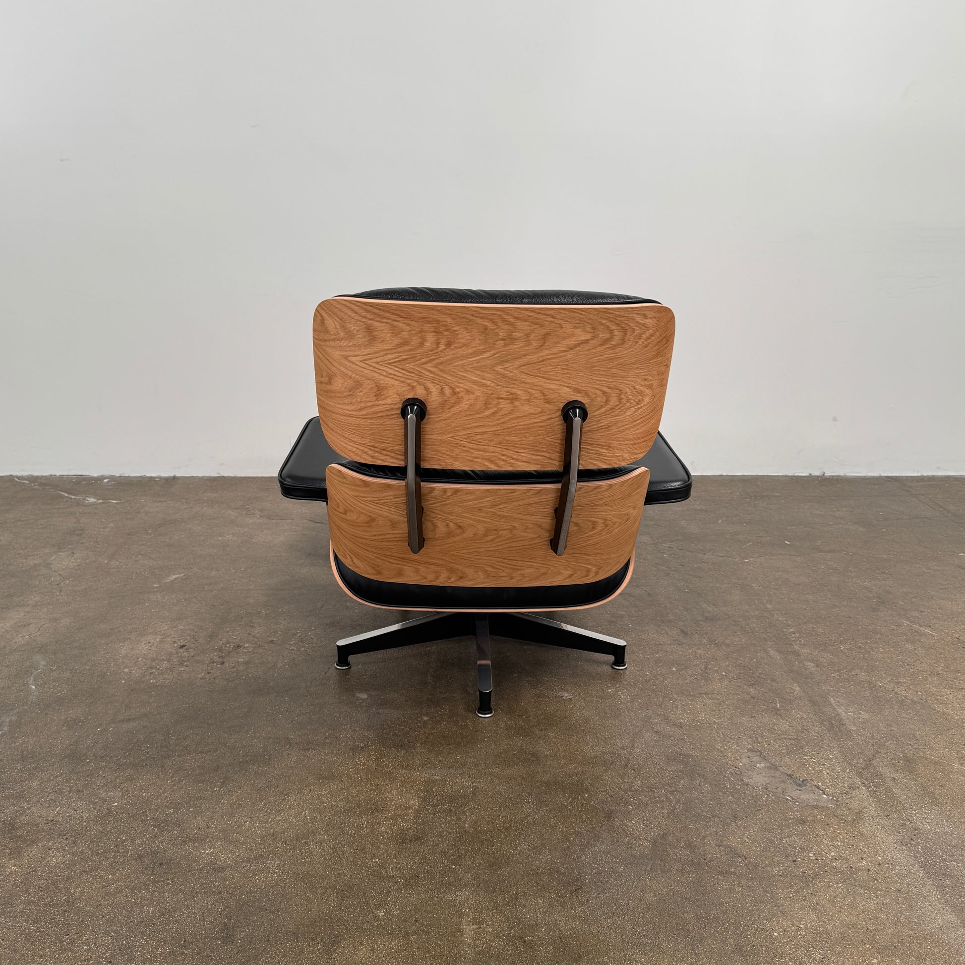 The Herman Miller Eames Lounge Chair & Ottoman (Copy) by Herman Miller, featuring black leather upholstery and modern design inspiration, is positioned on a brown concrete floor against a plain light-colored wall.