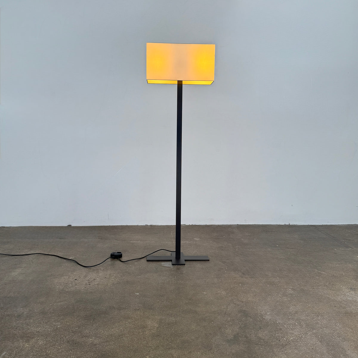 The Maxalto Leukon Tall Floor Lamp by Maxalto, featuring a minimalist design with a rectangular beige shade and a bronze nickel stand, stands on a bare concrete floor, plugged into an electrical outlet and casting ambient light throughout the space.