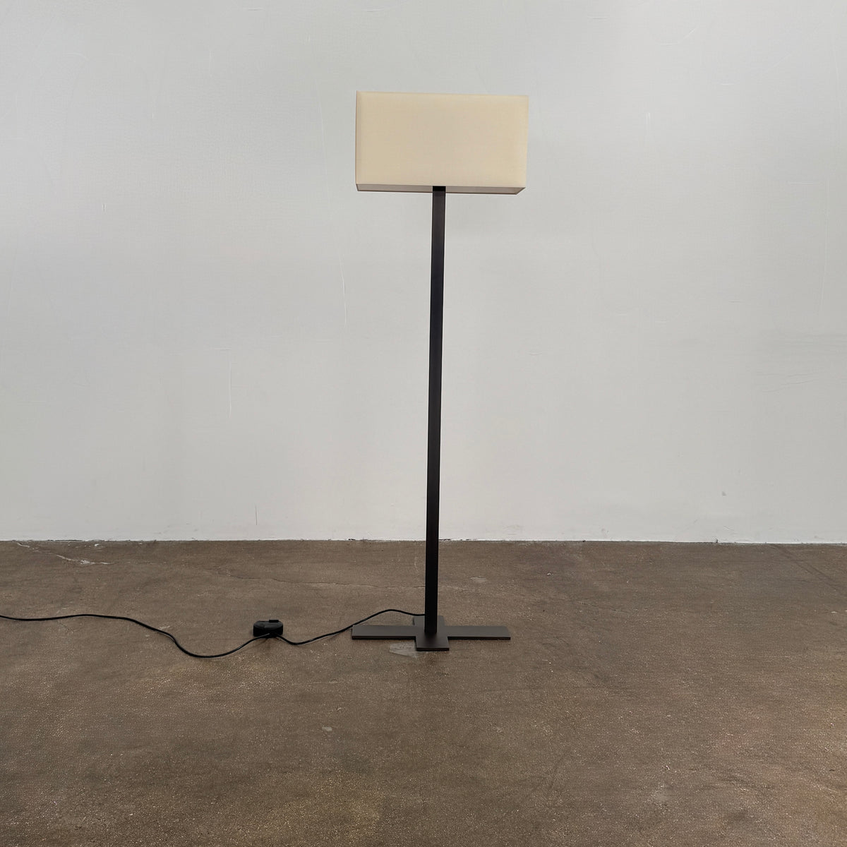 The Maxalto Leukon Tall Floor Lamp by Maxalto, featuring a minimalist design with a rectangular beige shade and a bronze nickel stand, stands on a bare concrete floor, plugged into an electrical outlet and casting ambient light throughout the space.