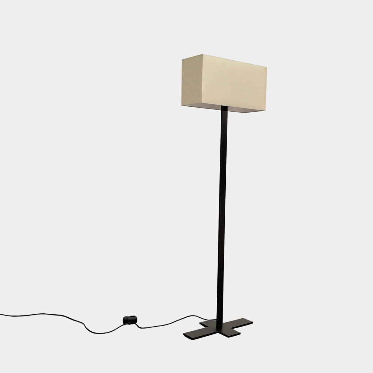 The Maxalto Leukon Tall Floor Lamp by Maxalto, featuring a minimalist design with a rectangular beige shade and a bronze nickel stand, stands on a bare concrete floor, plugged into an electrical outlet and casting ambient light throughout the space.