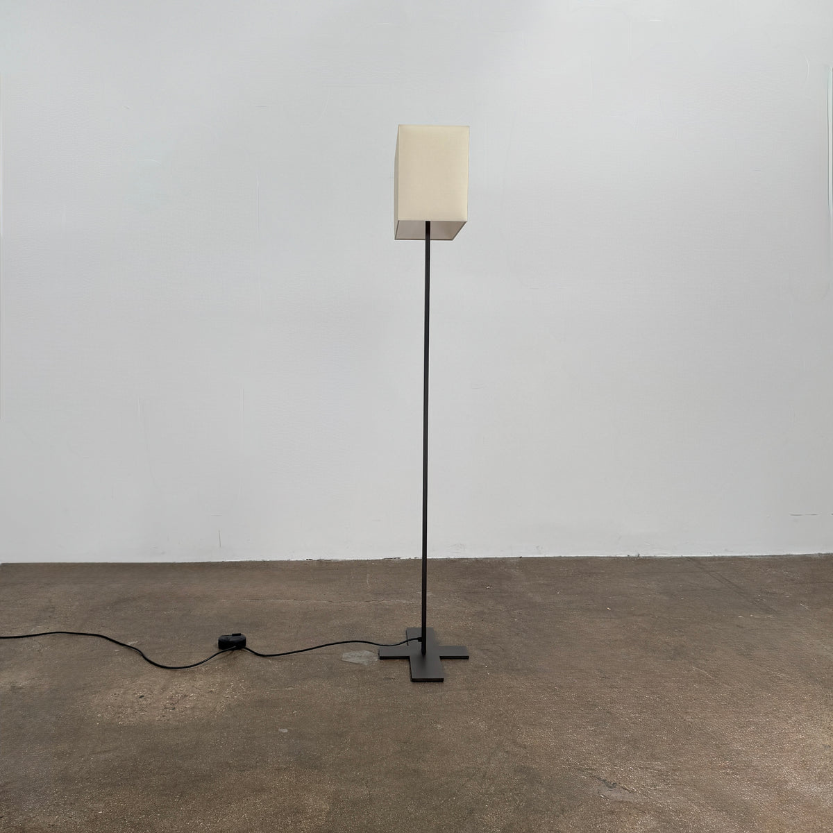 The Maxalto Leukon Tall Floor Lamp by Maxalto, featuring a minimalist design with a rectangular beige shade and a bronze nickel stand, stands on a bare concrete floor, plugged into an electrical outlet and casting ambient light throughout the space.