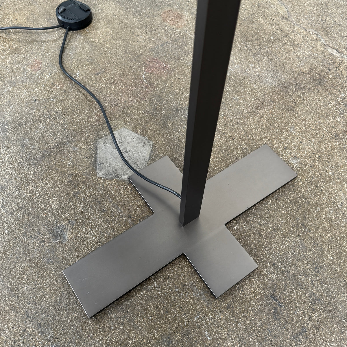 The Maxalto Leukon Tall Floor Lamp by Maxalto, featuring a minimalist design with a rectangular beige shade and a bronze nickel stand, stands on a bare concrete floor, plugged into an electrical outlet and casting ambient light throughout the space.
