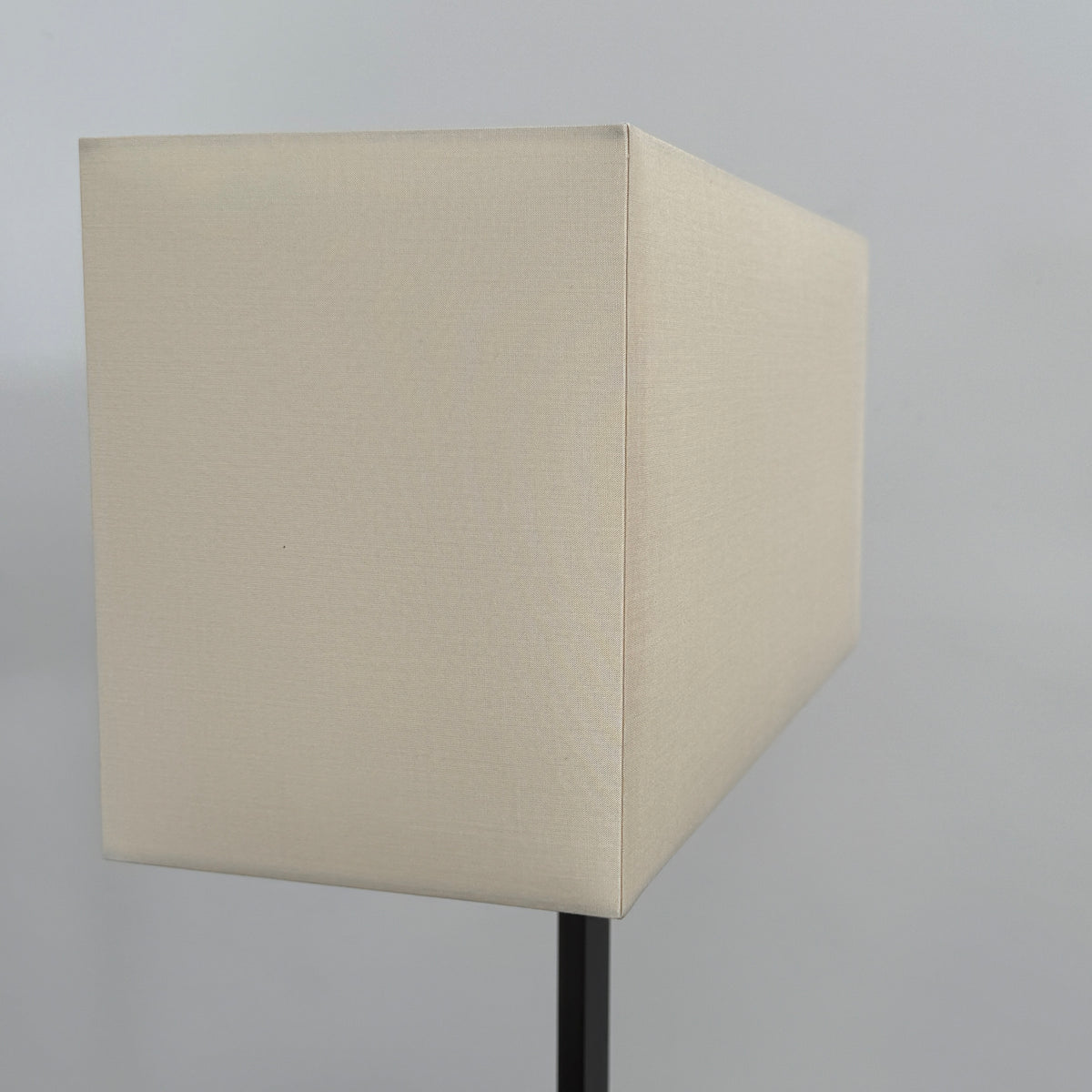 The Maxalto Leukon Tall Floor Lamp by Maxalto, featuring a minimalist design with a rectangular beige shade and a bronze nickel stand, stands on a bare concrete floor, plugged into an electrical outlet and casting ambient light throughout the space.
