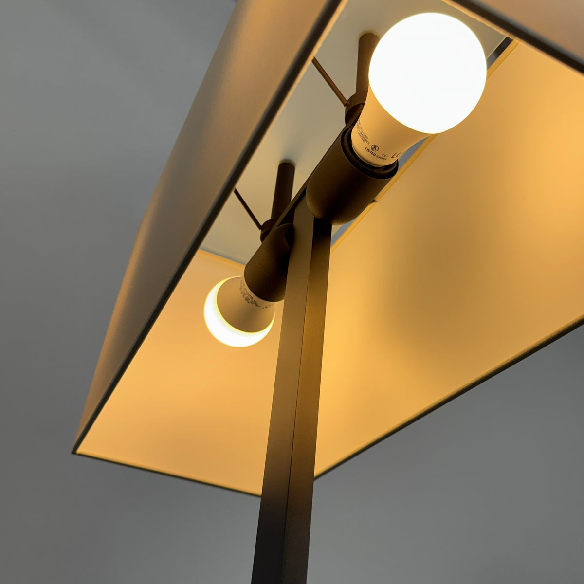 The Maxalto Leukon Tall Floor Lamp by Maxalto, featuring a minimalist design with a rectangular beige shade and a bronze nickel stand, stands on a bare concrete floor, plugged into an electrical outlet and casting ambient light throughout the space.