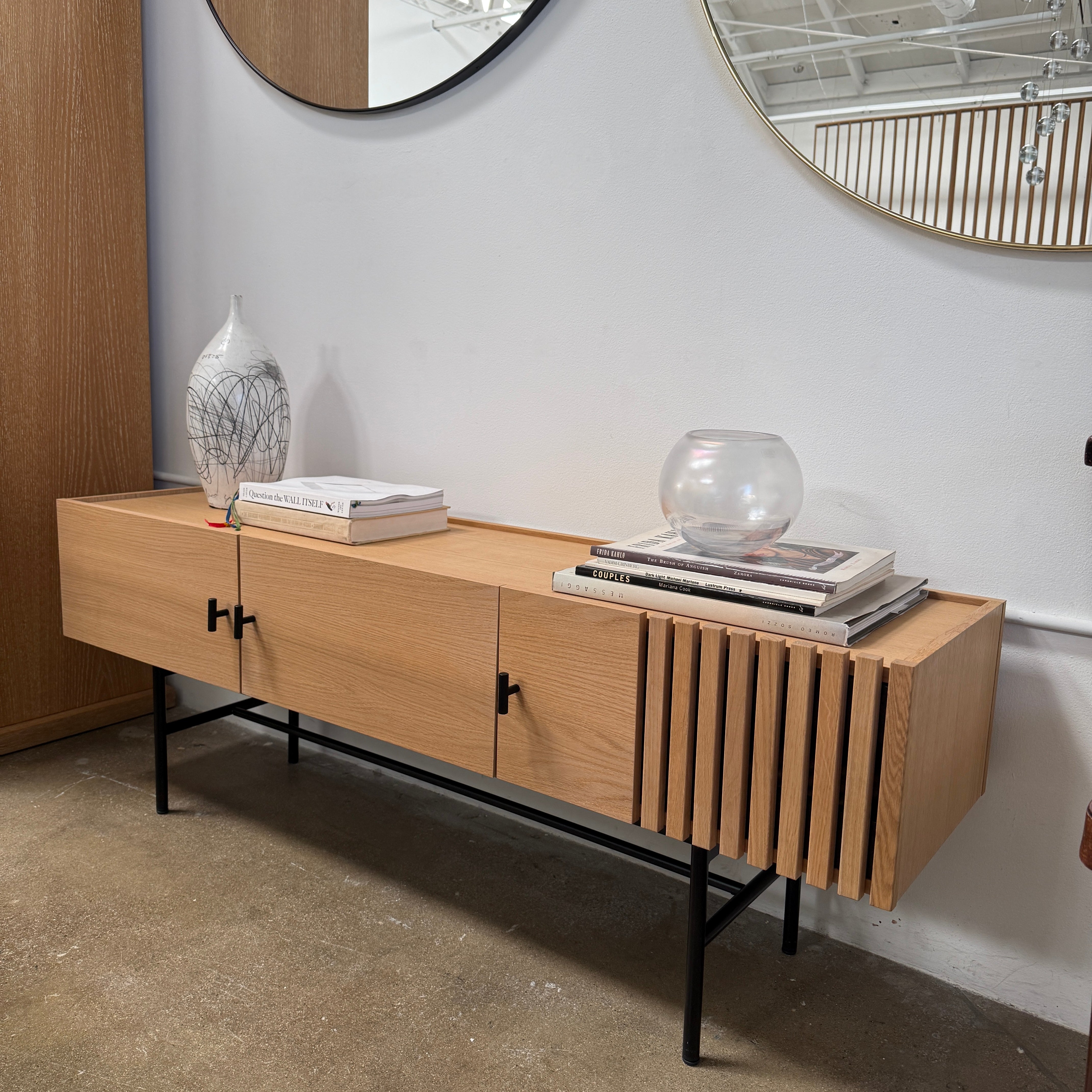 The Woud Array Low Sideboard in white pigmented oak by Modern Resale features three doors, black metal legs, and vertical slats on the right side, displayed against a plain white wall and concrete floor.