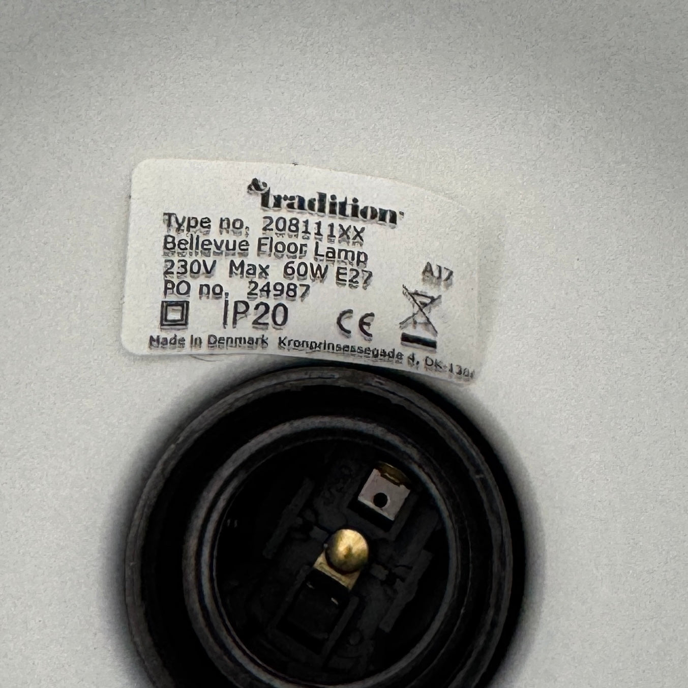 Close-up of a lamp label displaying model details—230V Max 60W E27, IP20—and manufacturer information above a black lamp socket, an elegant detail characteristic of the &Tradition Bellevue Floor Lamp AJ7 by &Tradition, a design that exemplifies Arne Jacobsen’s influential contribution to Scandinavian modernism.