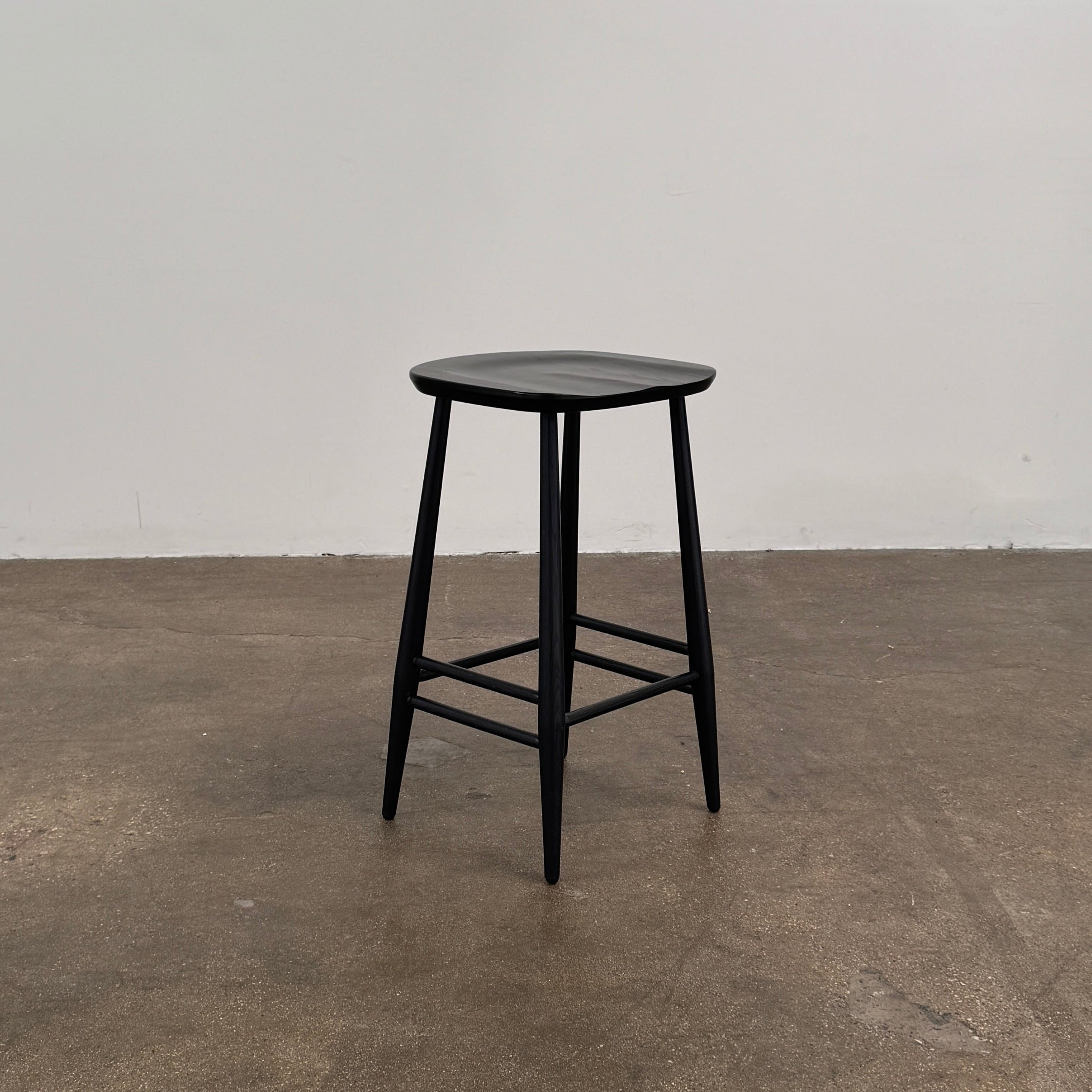 The Ercol Originals Counter Stool in Black Ash, crafted by Ercol, features four legs, a square contoured seat, and footrests on two sides. Set against a plain white background, this piece showcases the timeless craftsmanship of handmade British furniture.