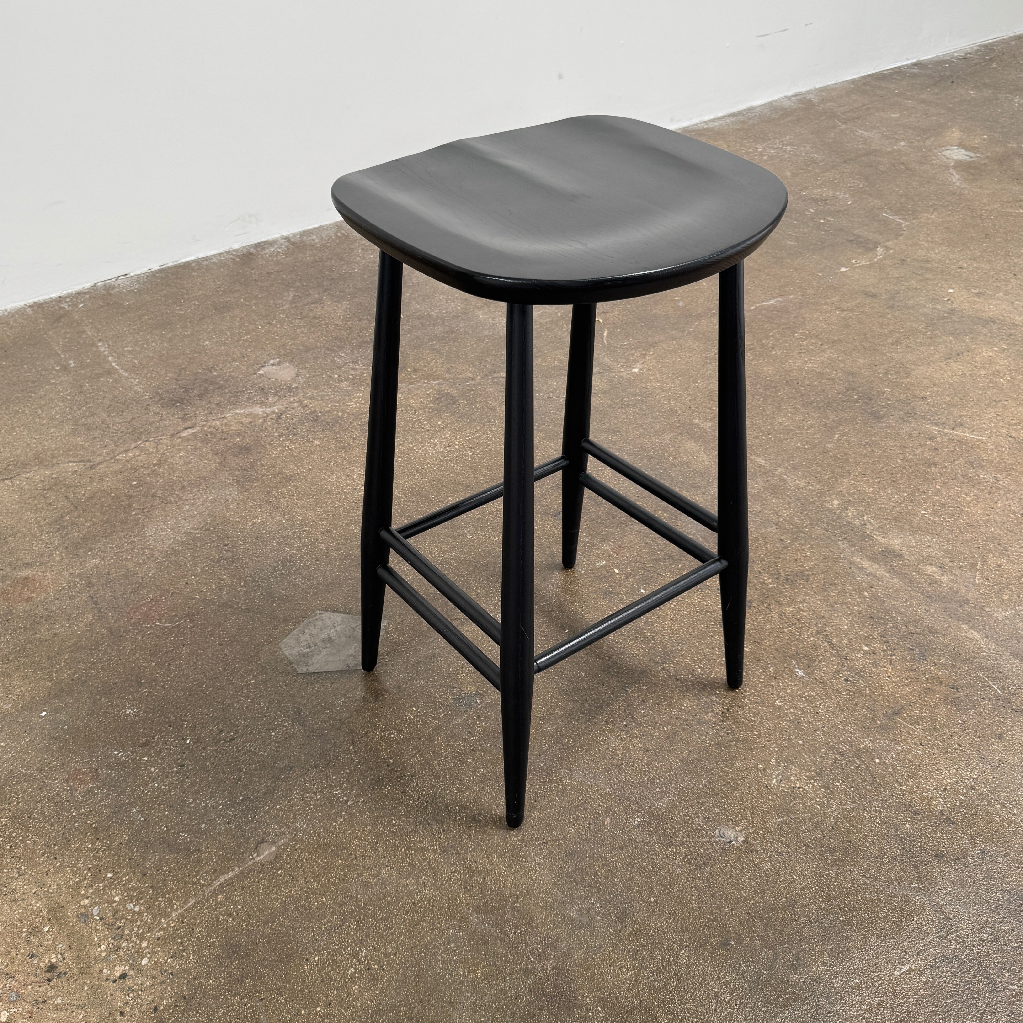 The Ercol Originals Counter Stool in Black Ash, crafted by Ercol, features four legs, a square contoured seat, and footrests on two sides. Set against a plain white background, this piece showcases the timeless craftsmanship of handmade British furniture.