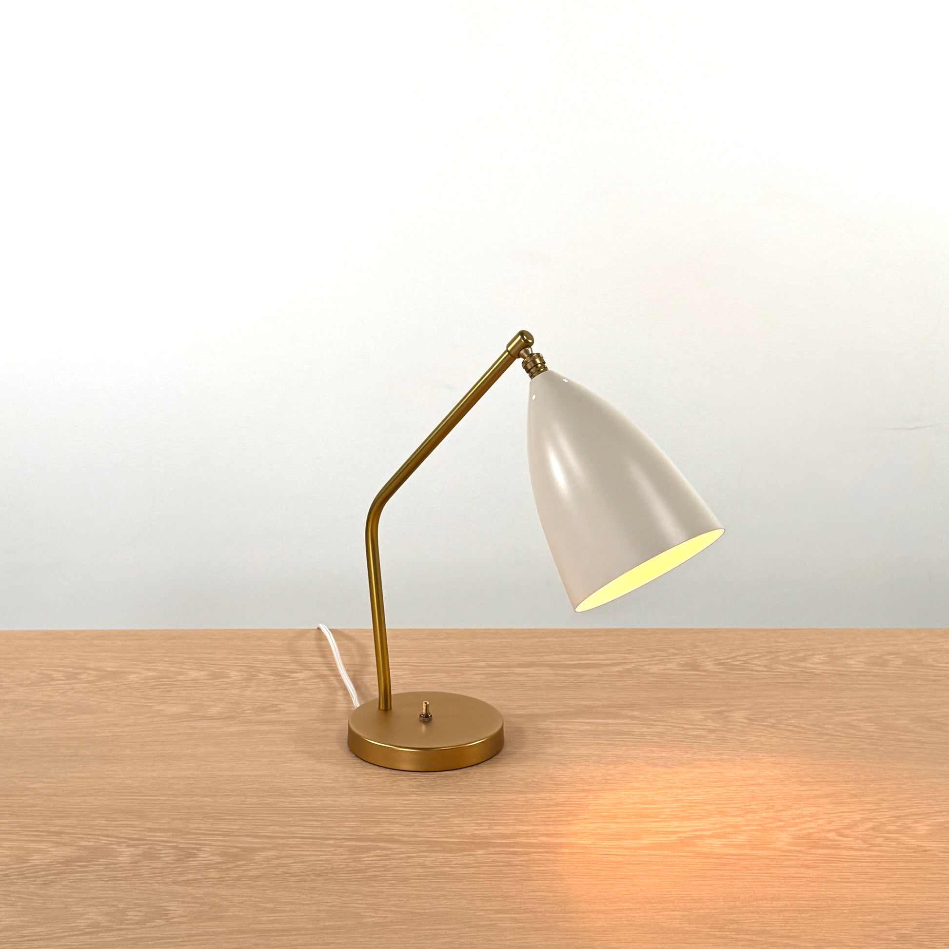 The Gubi Grashoppa Table Lamp in Alabaster White by Gubi sits on a light wood table against a plain white background, featuring its iconic brass design and white lampshade.
