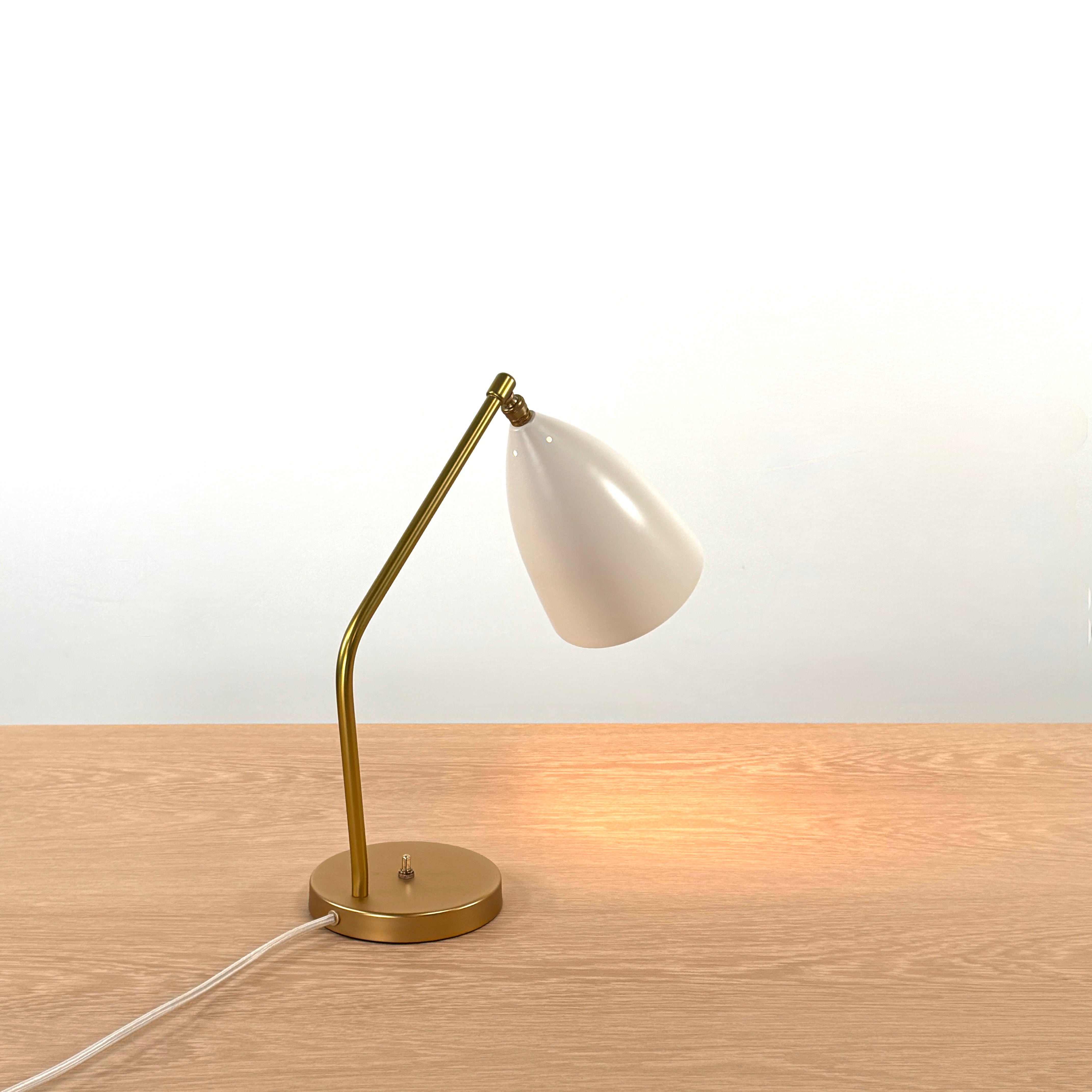 The Gubi Grashoppa Table Lamp in Alabaster White by Gubi sits on a light wood table against a plain white background, featuring its iconic brass design and white lampshade.