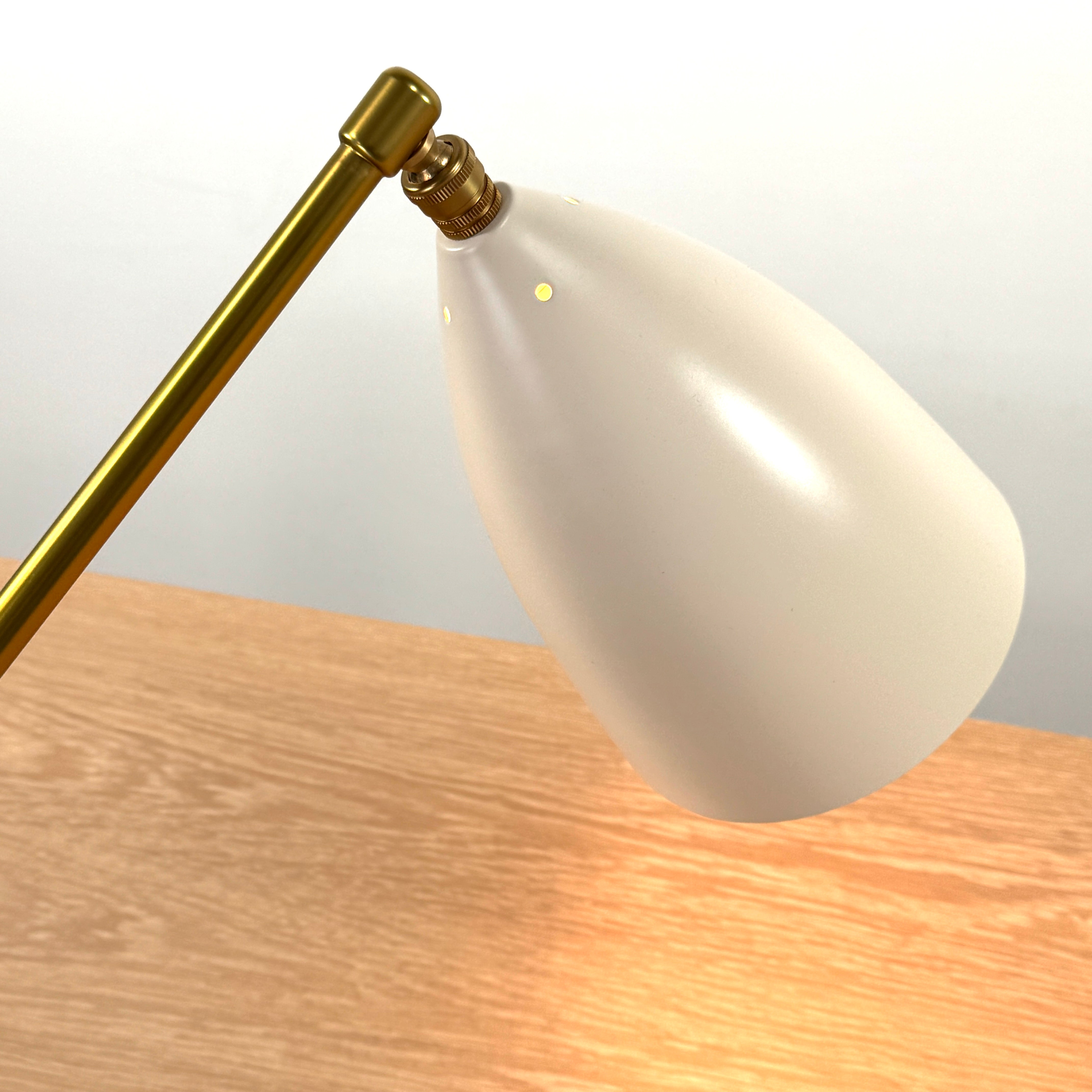 The Gubi Grashoppa Table Lamp in Alabaster White by Gubi sits on a light wood table against a plain white background, featuring its iconic brass design and white lampshade.