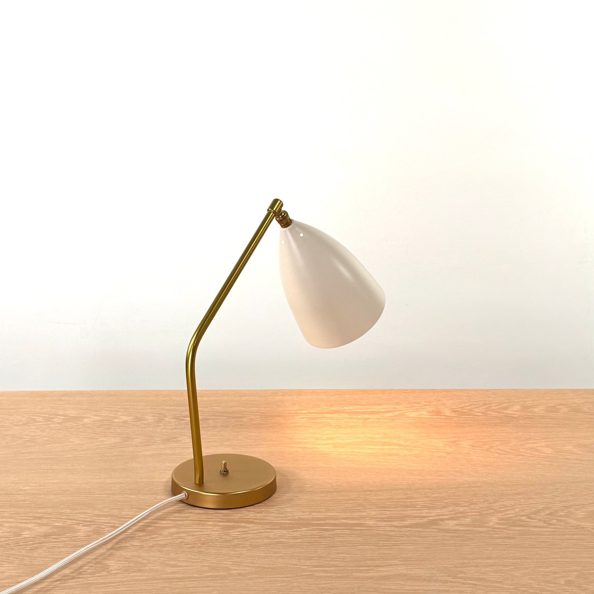 The Gubi Grashoppa Table Lamp in Alabaster White by Gubi sits on a light wood table against a plain white background, featuring its iconic brass design and white lampshade.