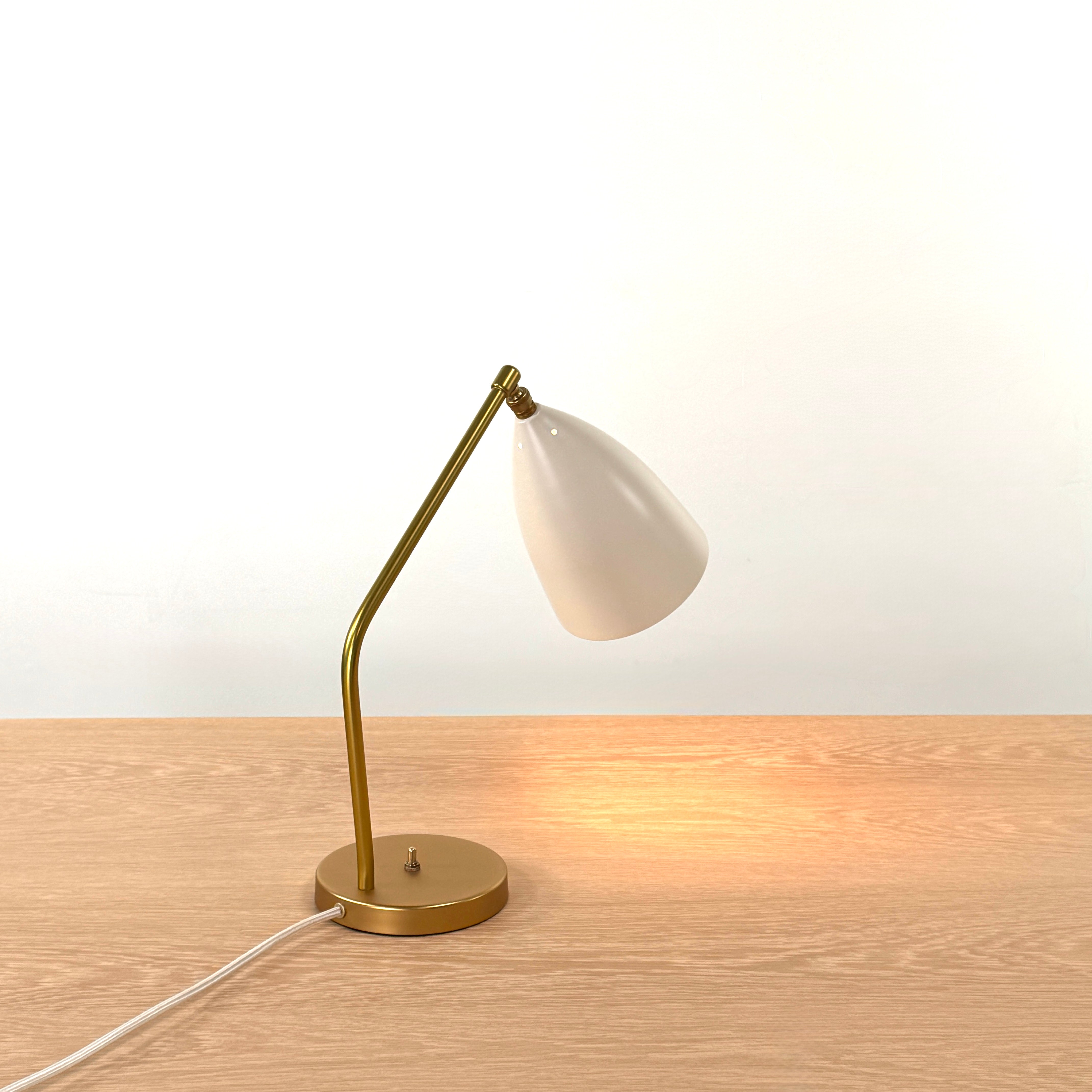 The Gubi Grashoppa Table Lamp in Alabaster White by Gubi sits on a light wood table against a plain white background, featuring its iconic brass design and white lampshade.