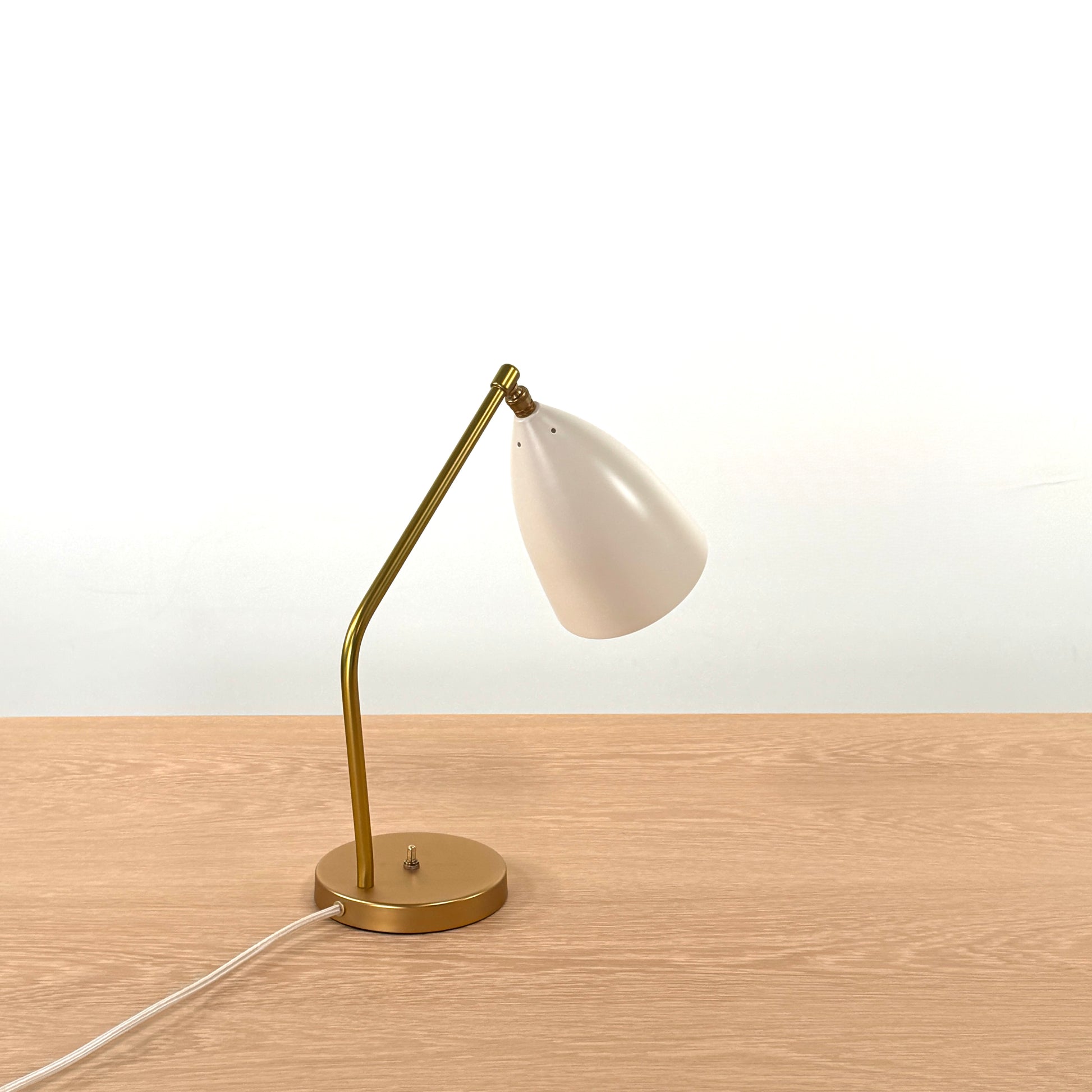 The Gubi Grashoppa Table Lamp in Alabaster White by Gubi sits on a light wood table against a plain white background, featuring its iconic brass design and white lampshade.