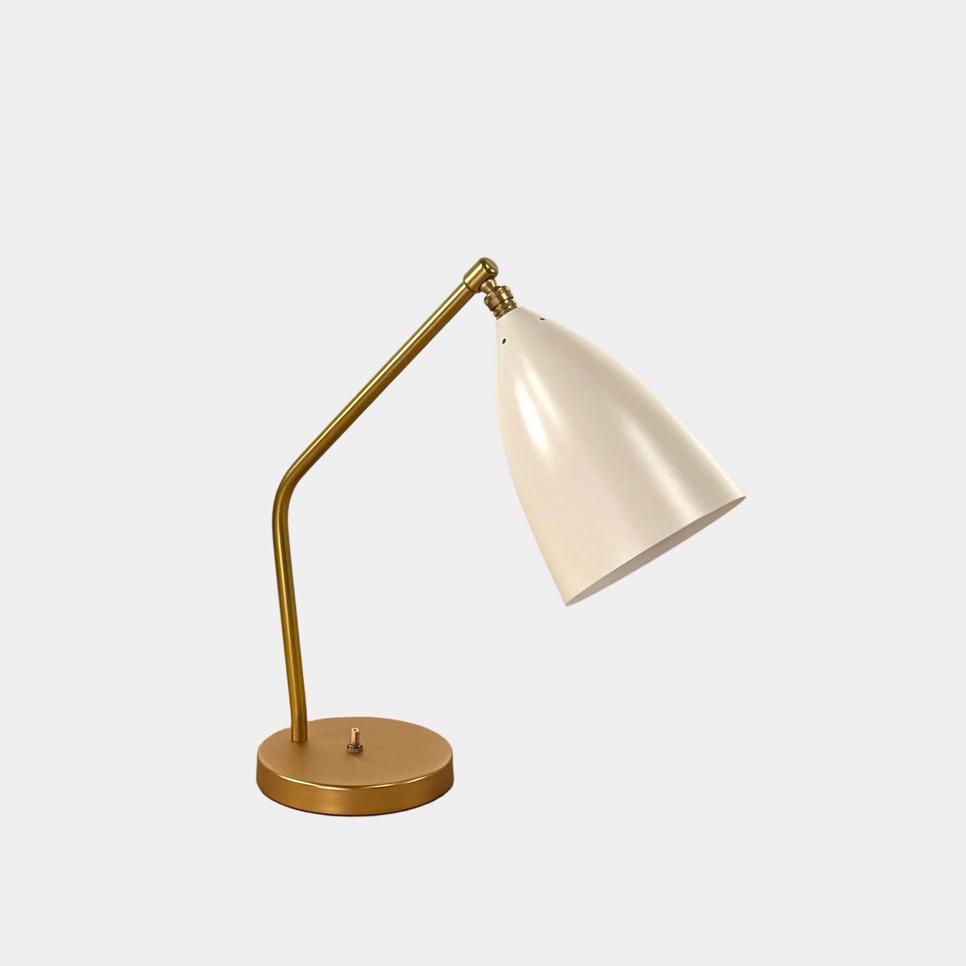 The Gubi Grashoppa Table Lamp in Alabaster White by Gubi sits on a light wood table against a plain white background, featuring its iconic brass design and white lampshade.