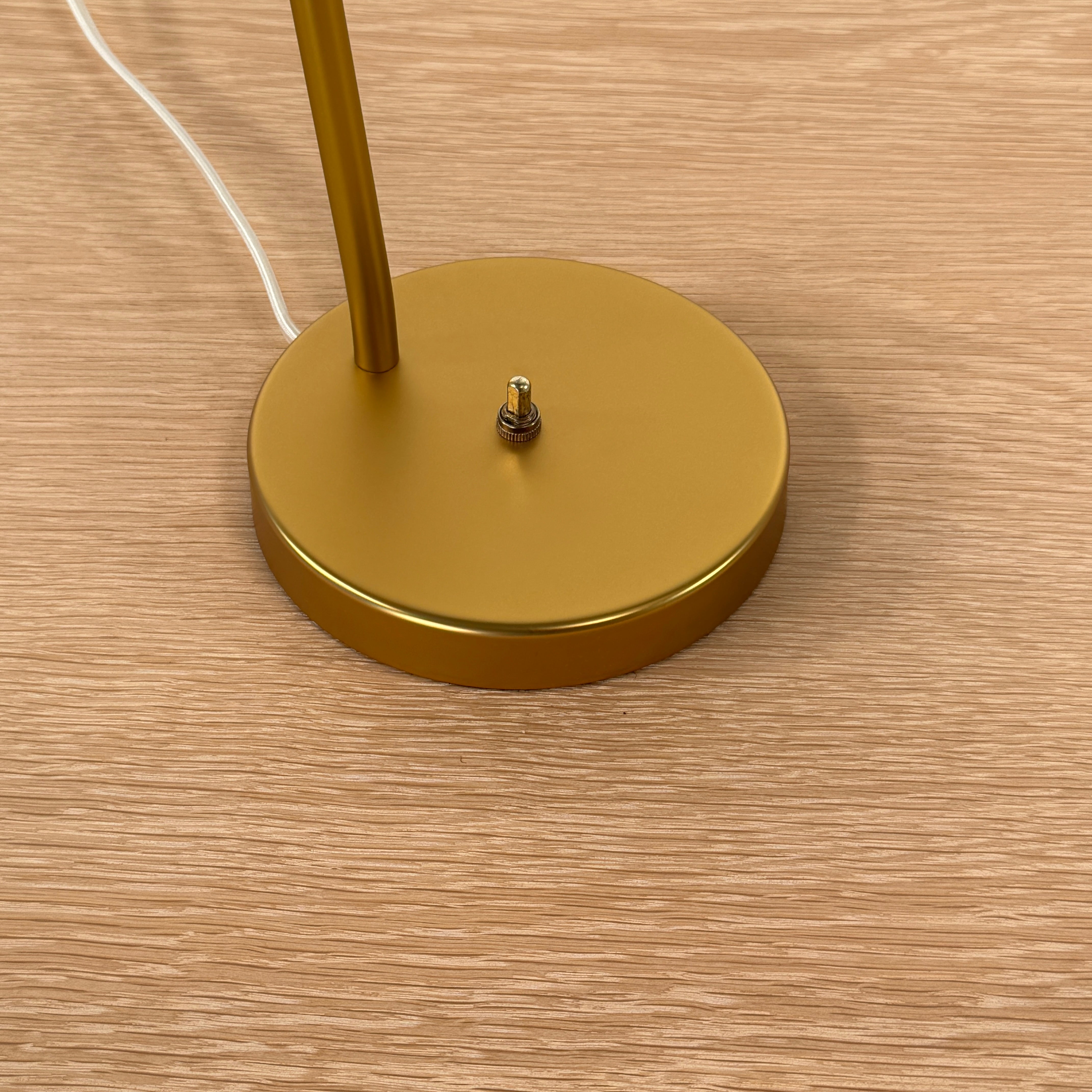 The Gubi Grashoppa Table Lamp in Alabaster White by Gubi sits on a light wood table against a plain white background, featuring its iconic brass design and white lampshade.