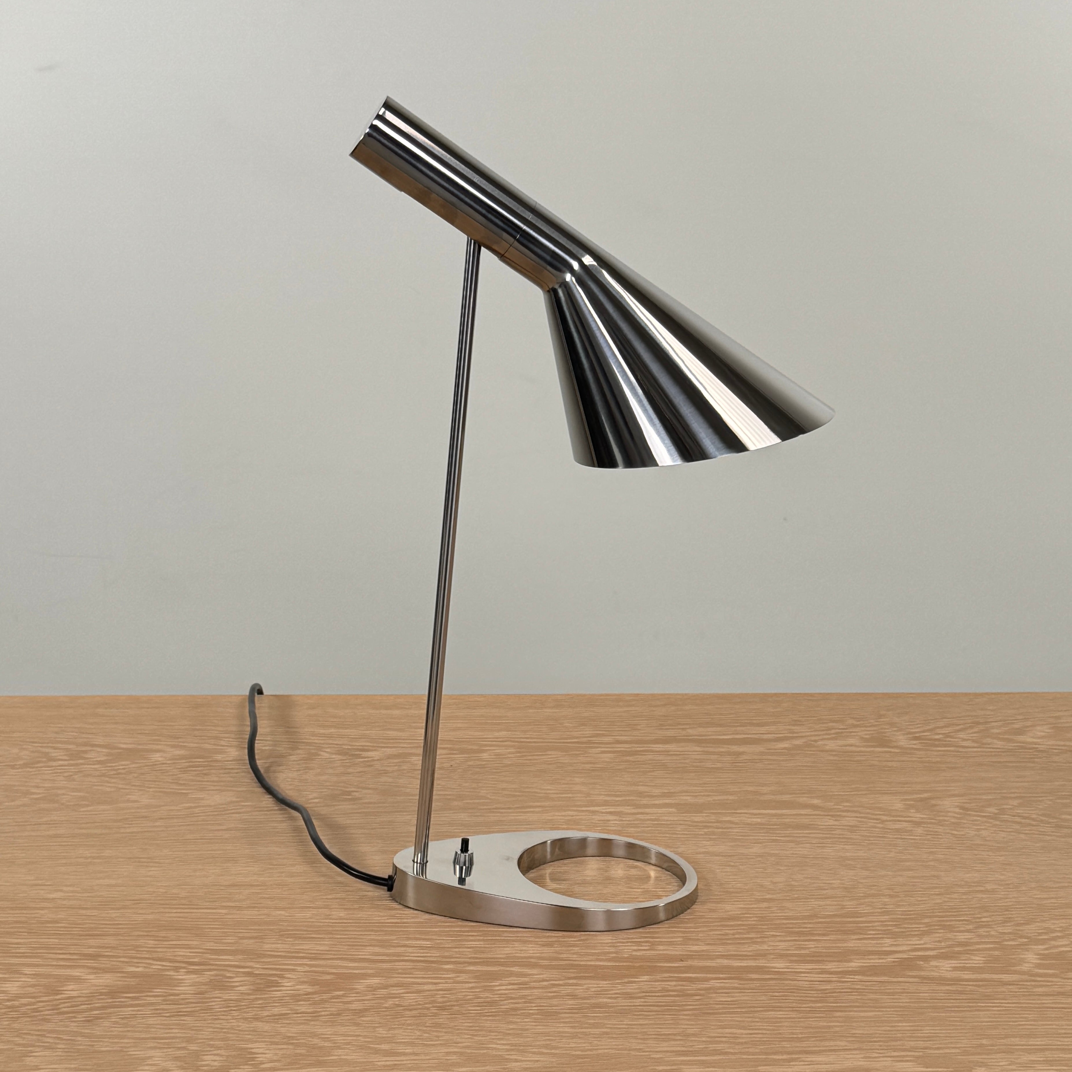 The Louis Poulsen AJ Table Lamp, featuring a modern metallic finish and slanted shade, rests on a wooden surface and illuminates the desk.